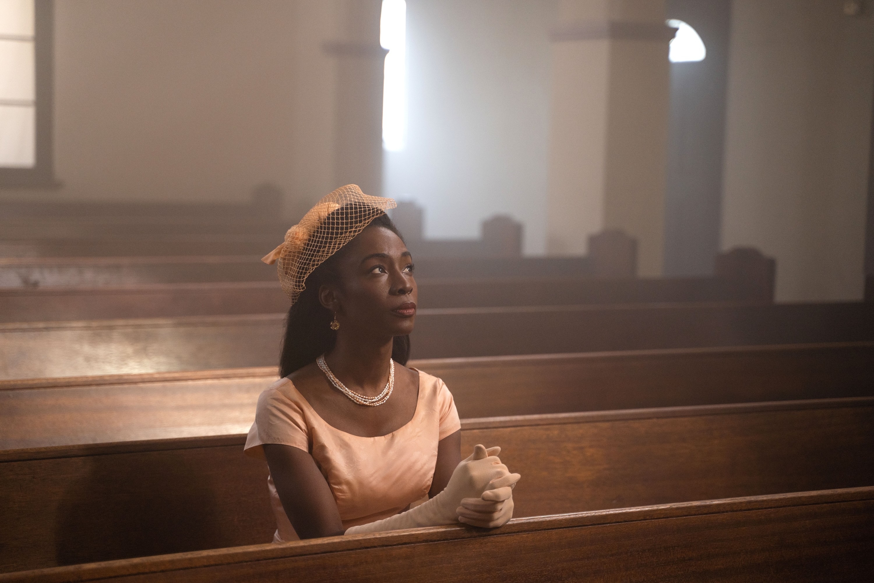 A woman in vintage attire sits alone in a church pew, wearing a pearl necklace, gloves, and a netted hat, gazing thoughtfully