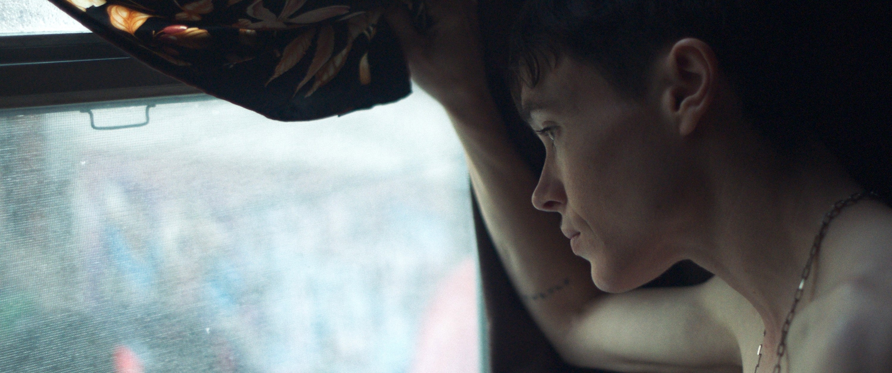 Elliot Page looks out a window, partially hidden by a floral curtain, appearing contemplative