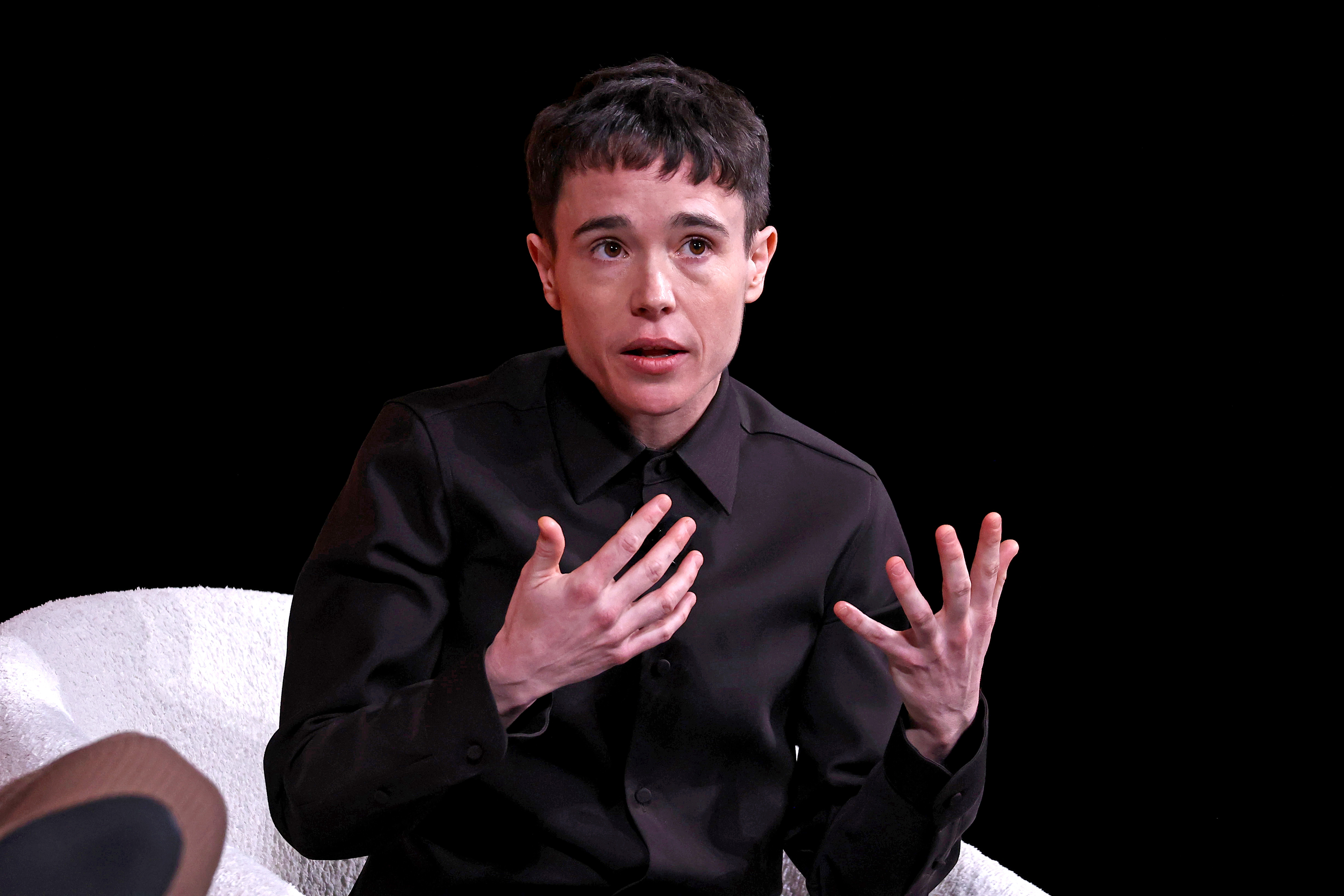 Elliot Page speaking on stage, wearing a black button-up shirt, with expressive hand gestures