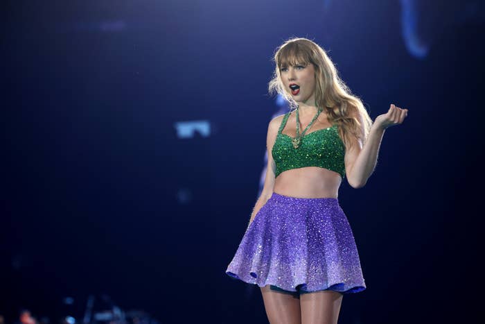 Taylor Swift performs on stage wearing a green sequin crop top and a purple high-waisted skirt. She stands with her hand raised, looking out at the audience