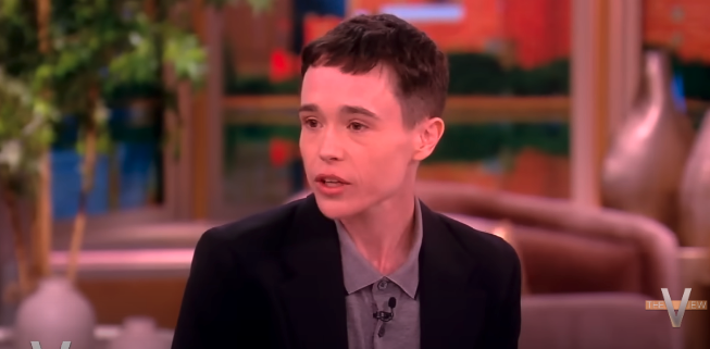 Elliot Page speaks during an interview on "The View," wearing a dark blazer over a gray shirt. Background features blurred studio decor