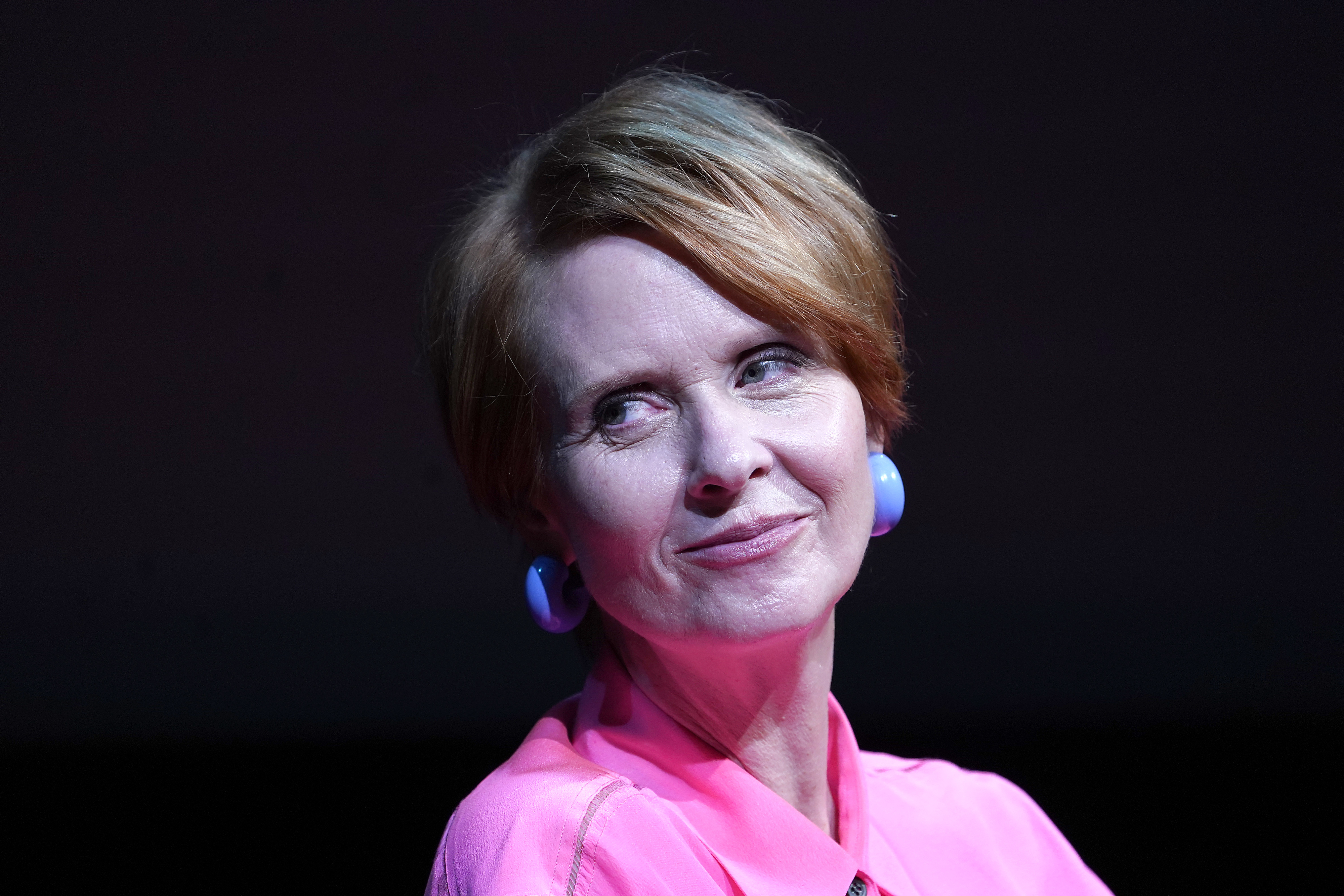 Cynthia Nixon wears a collared shirt and bold round earrings while giving a slight smile, featured in a close-up shot