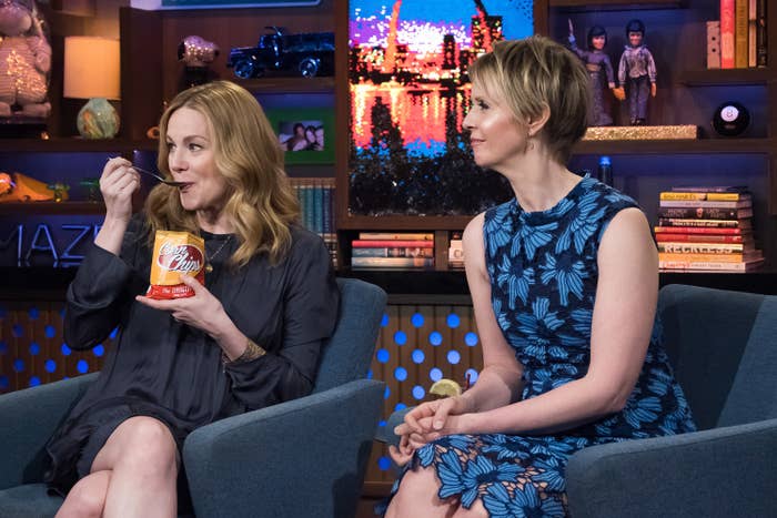 Elizabeth Reaser in casual attire, eating from a can, and Cynthia Nixon in a floral dress, sitting on a talk show set