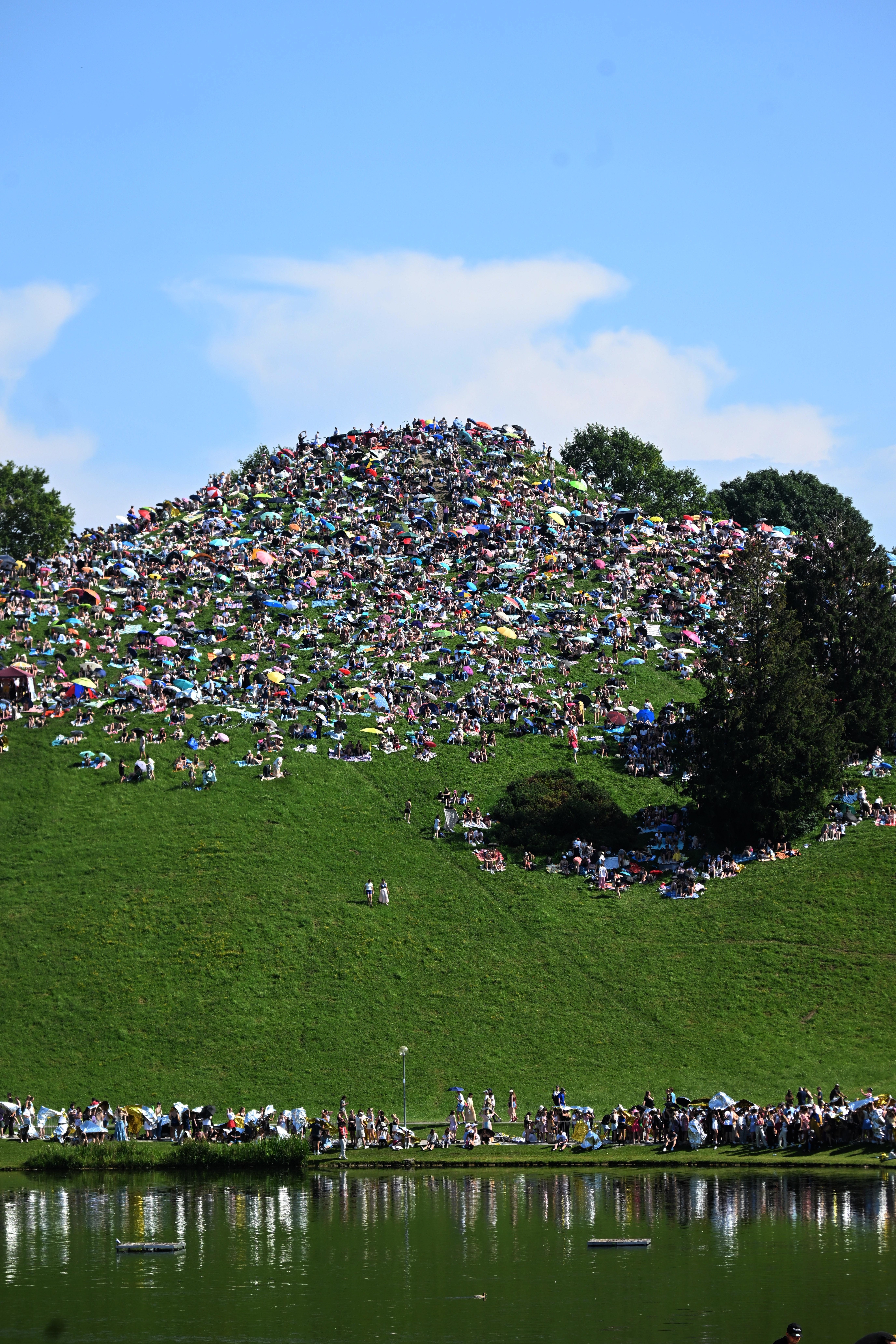 A large crowd of people cover a grassy hill and gather around a pond, likely at a festival or outdoor event