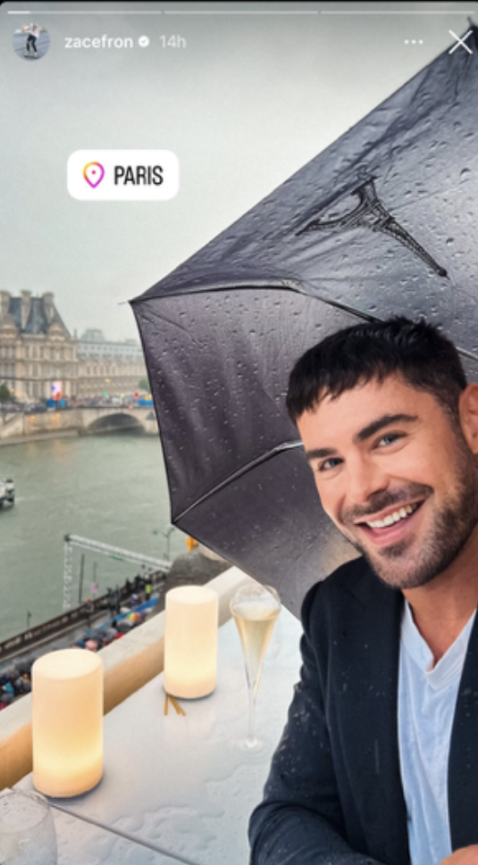 Zac Efron smiles under an umbrella on a rainy Paris balcony, overlooking the Seine River with the Louvre in the background