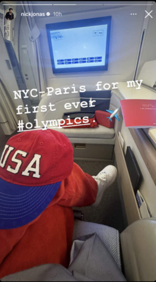 Nick Jonas in a plane seat, wearing a red USA cap, with text: "NYC-Paris for my first ever #olympics."