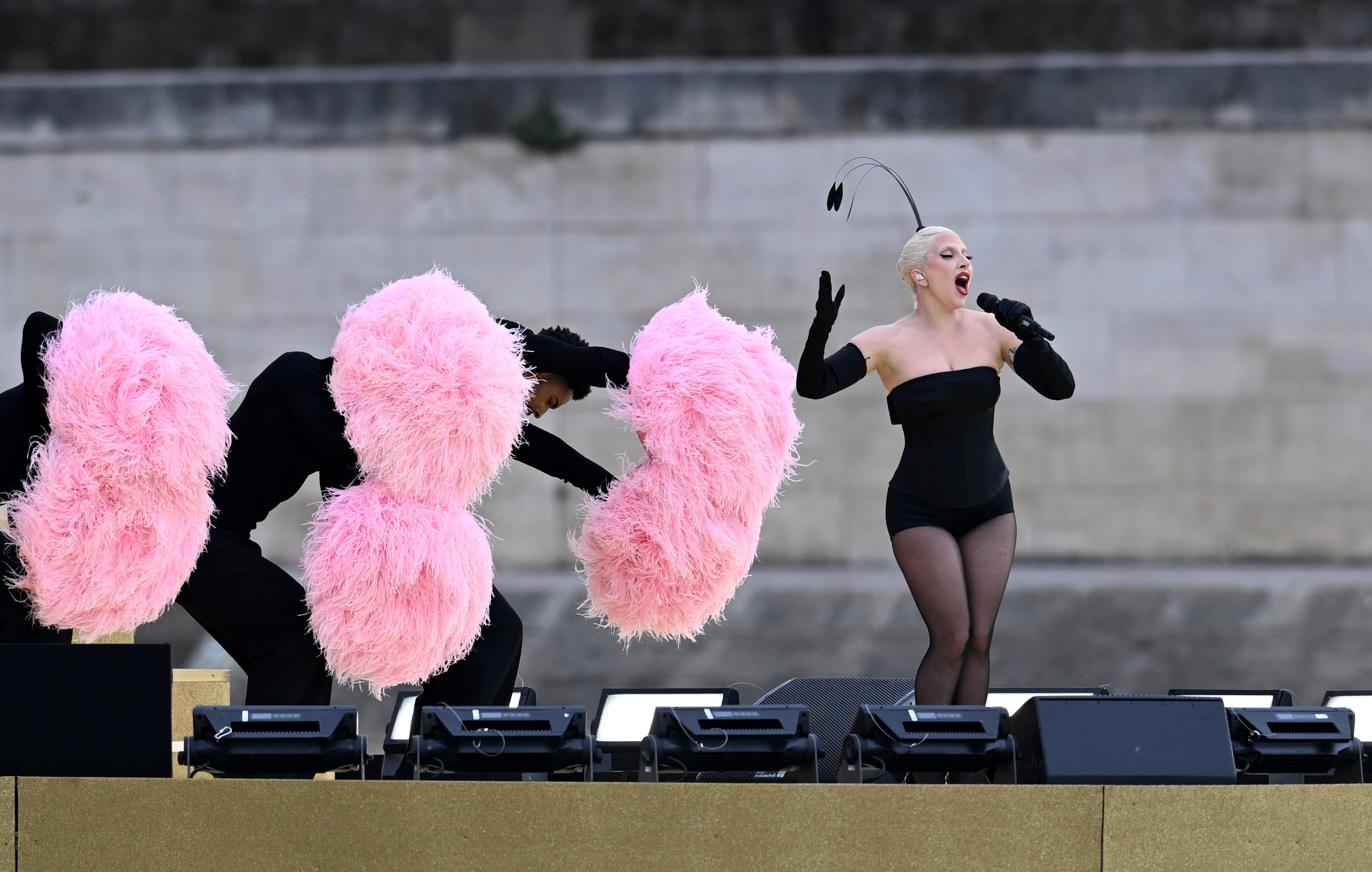 Lady Gaga performs in a black leotard and opera gloves, singing into a microphone, while backup dancers with large pink feather props dance beside her