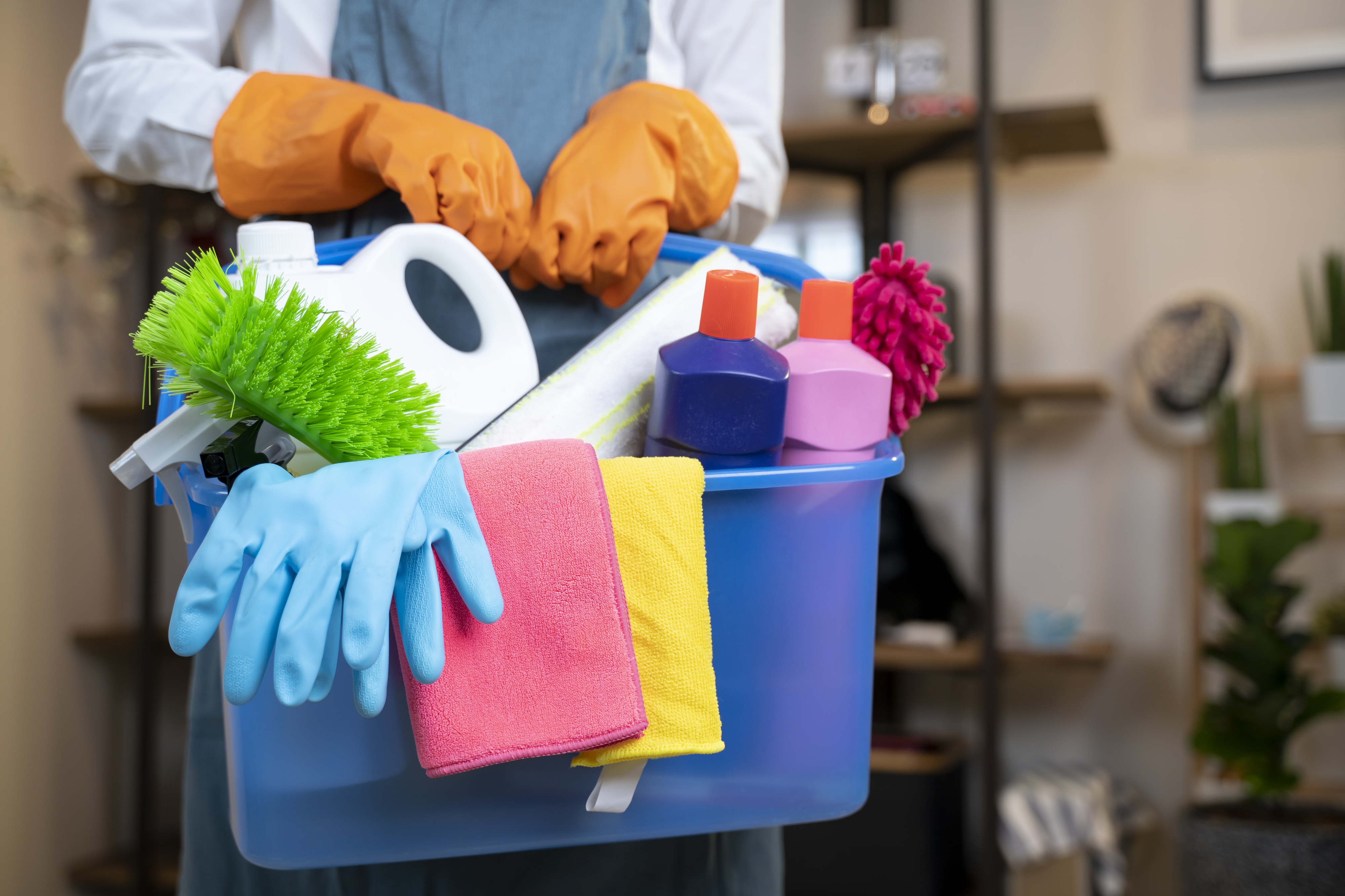 A person wearing orange gloves holds a blue bucket filled with cleaning supplies, including brushes, gloves, a spray bottle, and cloths