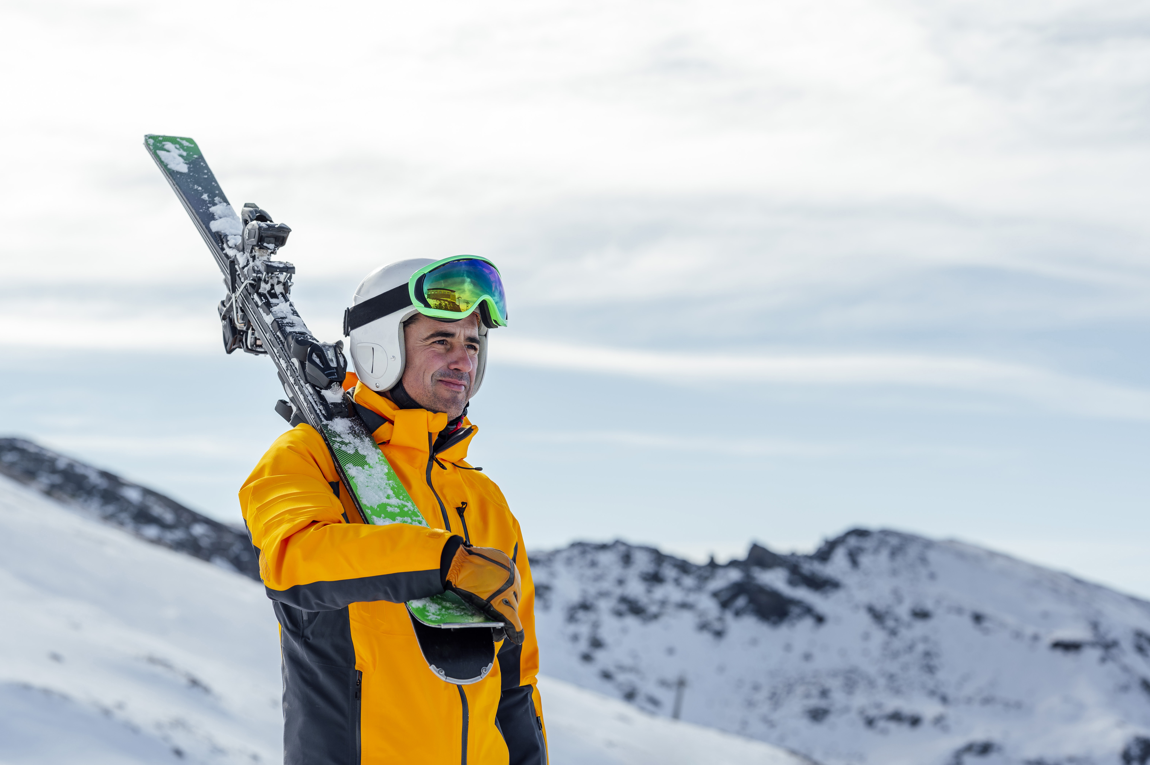 A man wearing a helmet, ski jacket, and goggles holds skis over his shoulder while standing on a snowy mountain