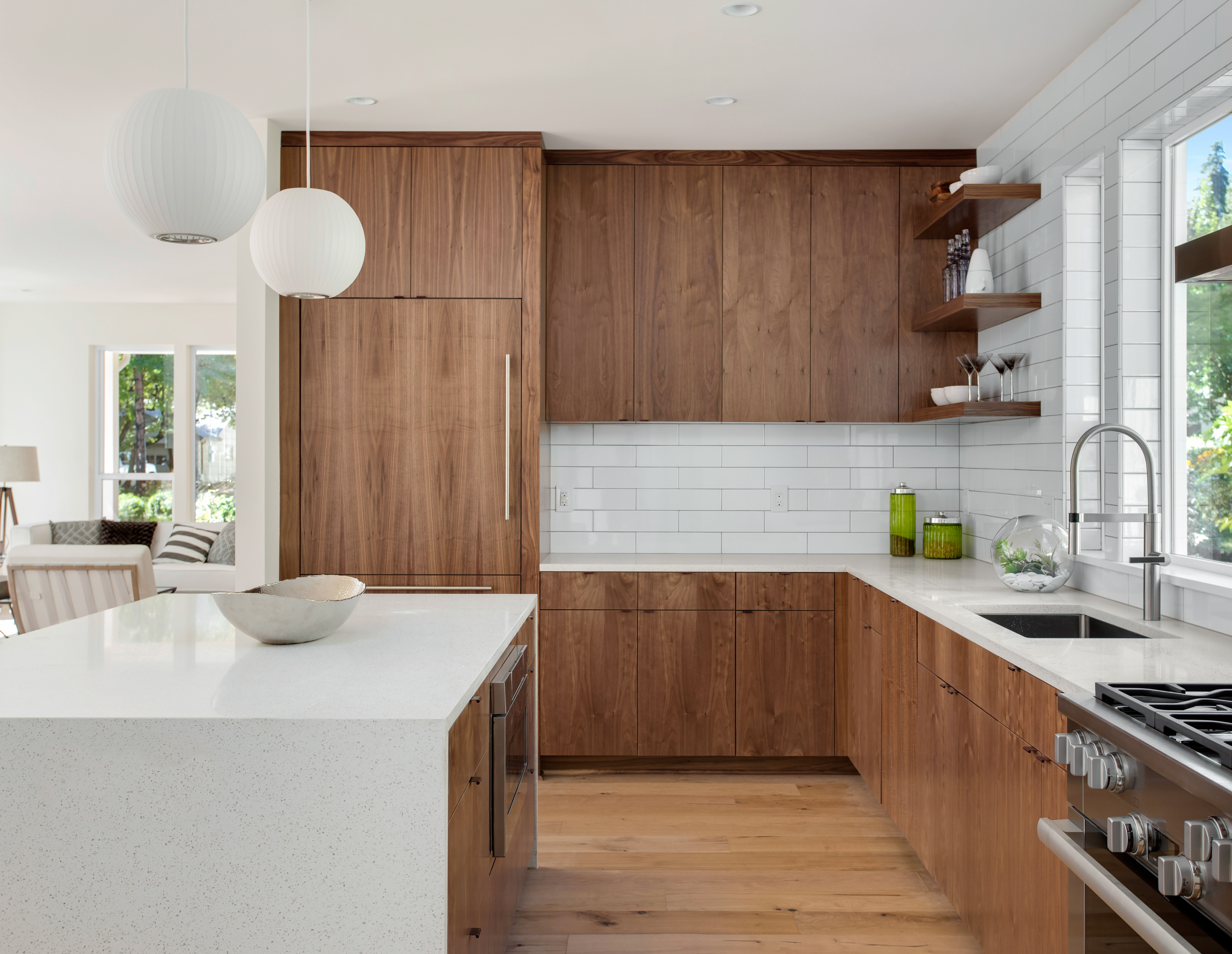 Modern kitchen with wooden cabinets, white countertops, stainless steel appliances, and large pendant lights over an island. Sunlight filters through windows