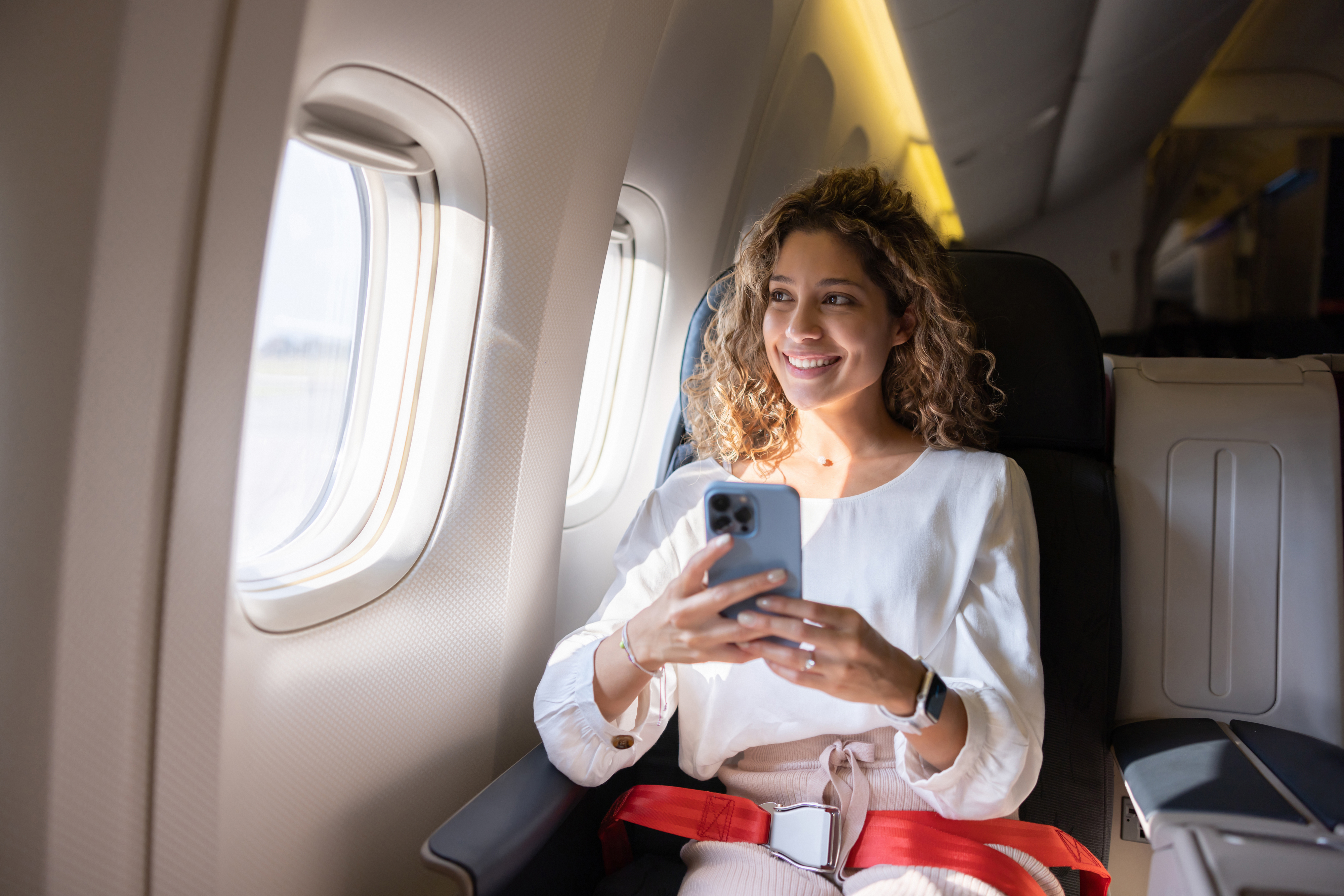 A woman in business class on an airplane, smiling while looking at her phone. She is wearing business casual attire. The image is part of a Work &amp;amp; Money article