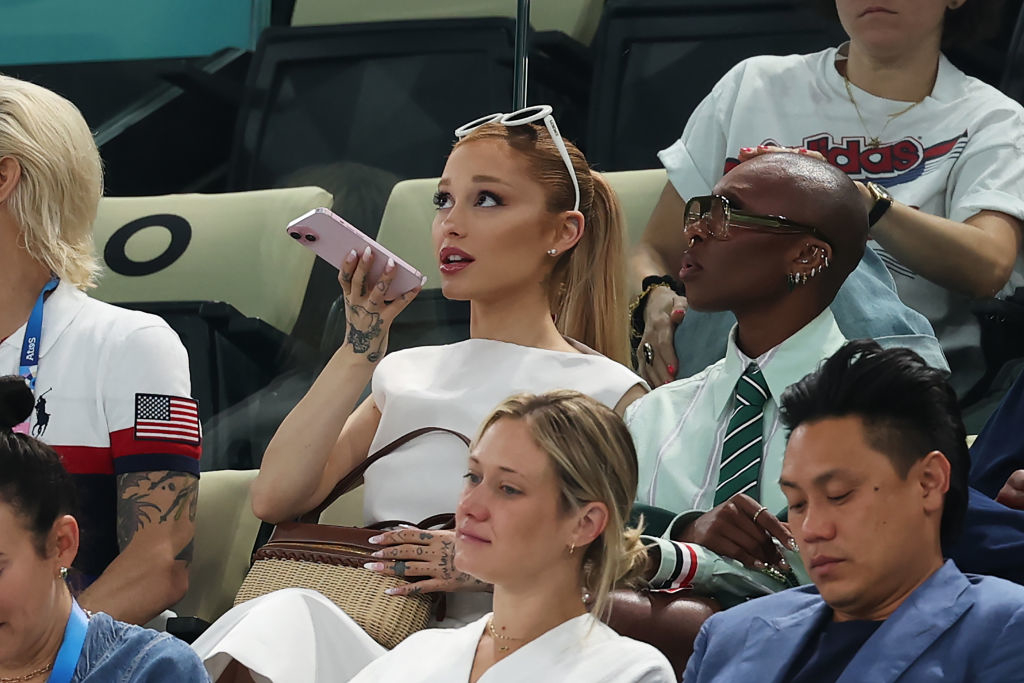 Ariana Grande sits in a stadium, dressed in a chic white outfit and holding a phone to her mouth. She is surrounded by other attendees, watching intently