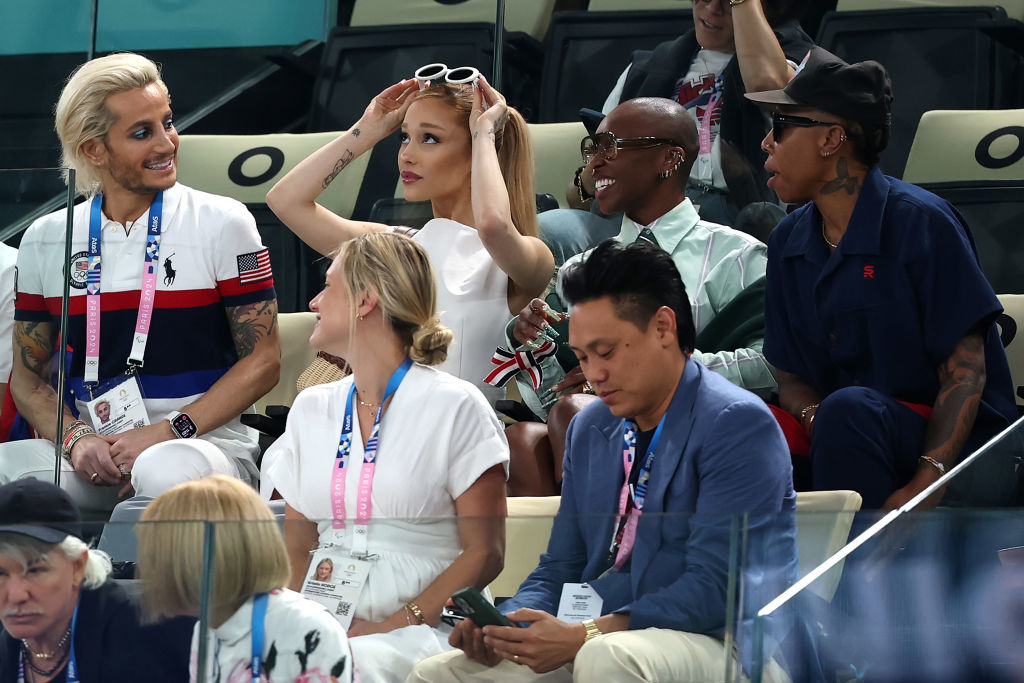Frankie Grande, Ariana Grande, and others sit in a sports arena, mingling and smiling. Ariana adjusts her sunglasses while Frankie chats with someone nearby