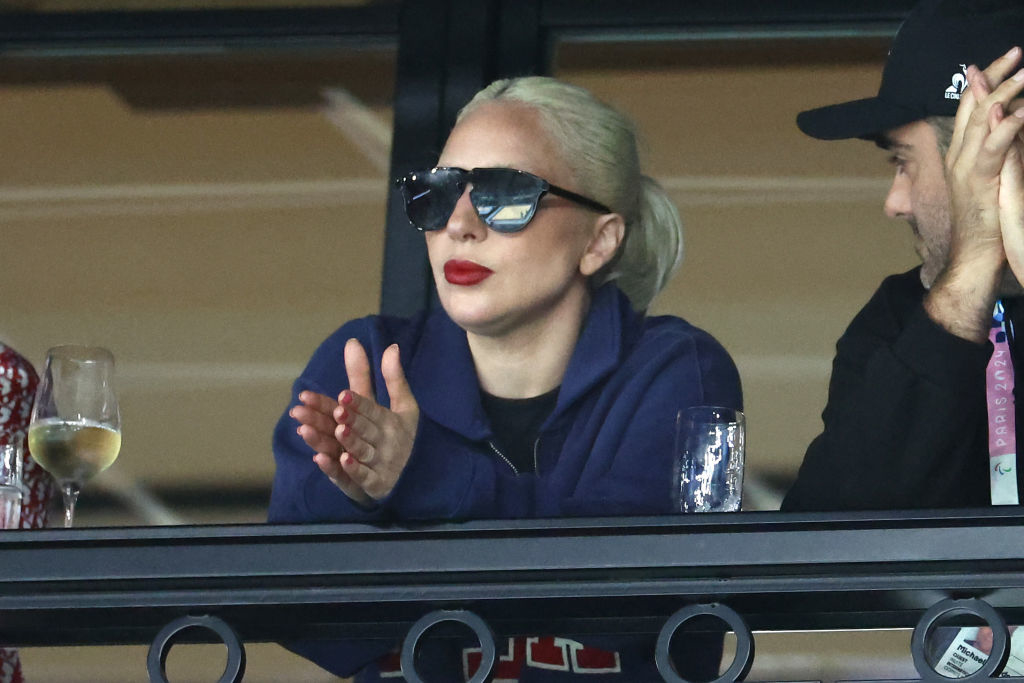 Lady Gaga, in a casual outfit with large sunglasses and red lipstick, claps while seated next to a man at an event