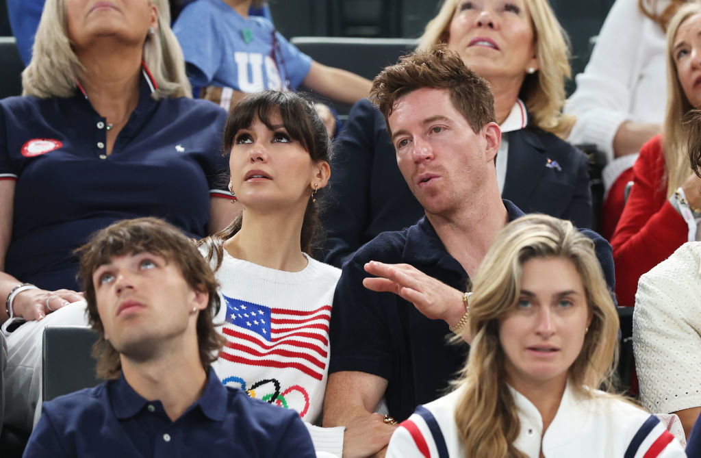 Shaun White, Nina Dobrev, and others sit in stadium seats, watching an event. Nina wears a white sweater with an American flag and Olympic rings