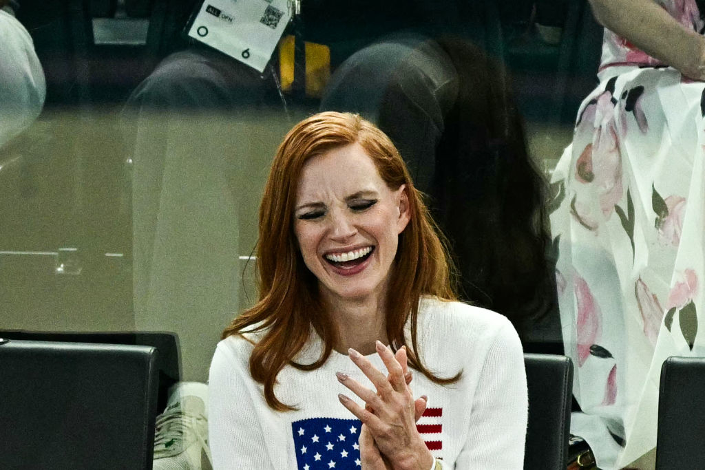 Jessica Chastain laughing, wearing a white sweater with an American flag design, attending an event