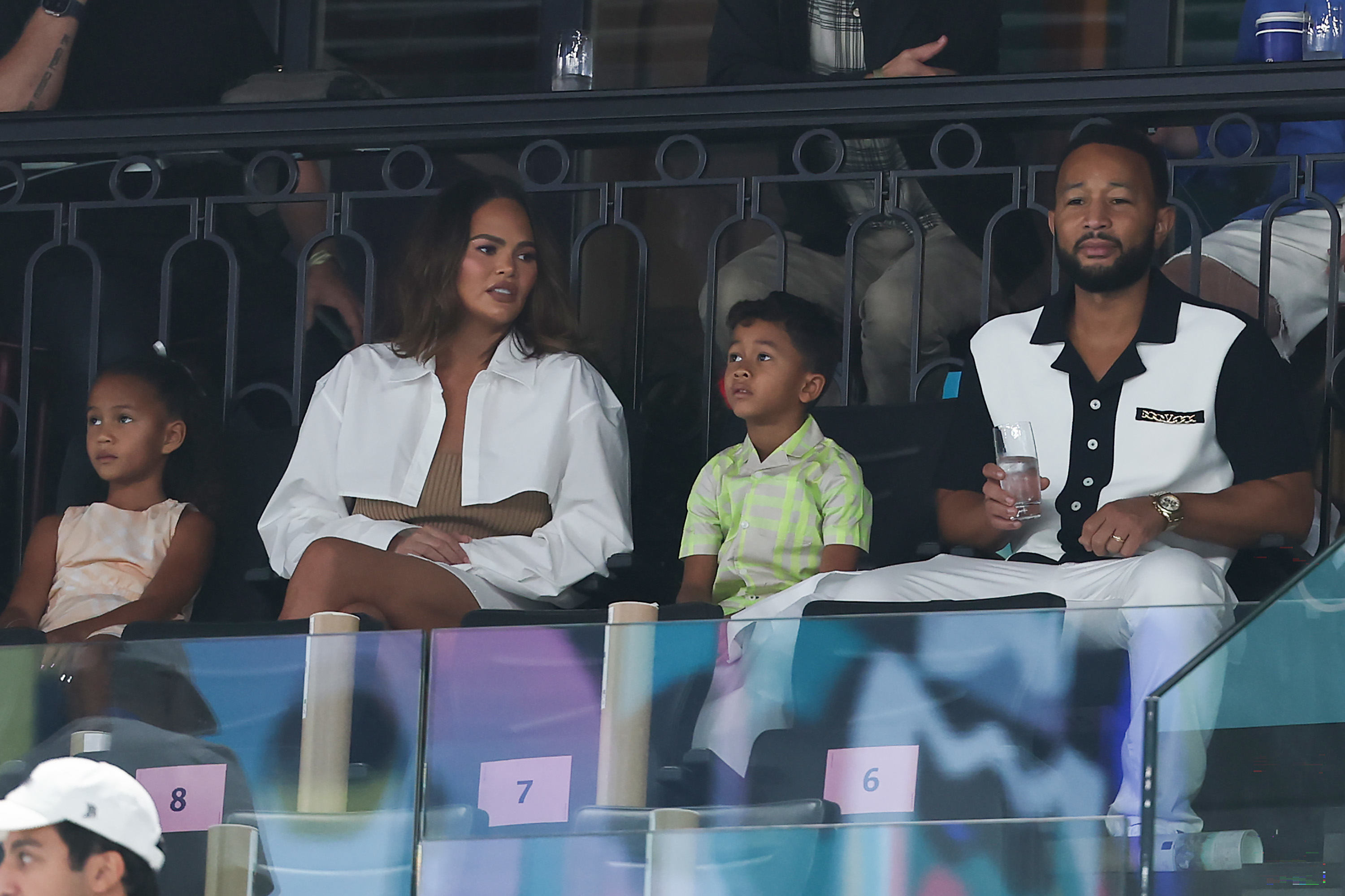 Chrissy Teigen and John Legend sit with their children at a sporting event. Chrissy wears a white jacket. John wears a white and black outfit