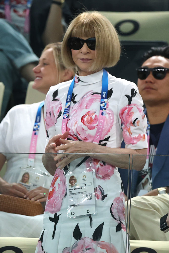 Anna Wintour in a floral dress and sunglasses at an event, wearing a lanyard and nametag. People are seated behind her