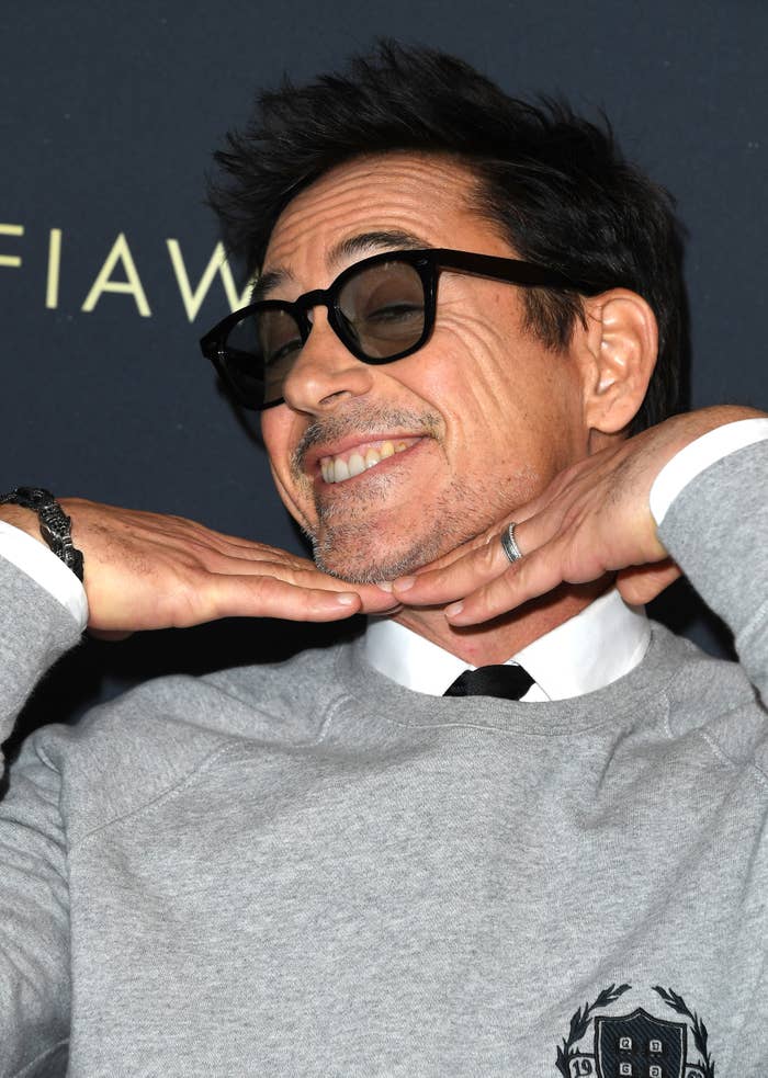 Robert Downey Jr. smiles with his hands under his chin, wearing a sweater, shirt, tie, and black-rimmed glasses