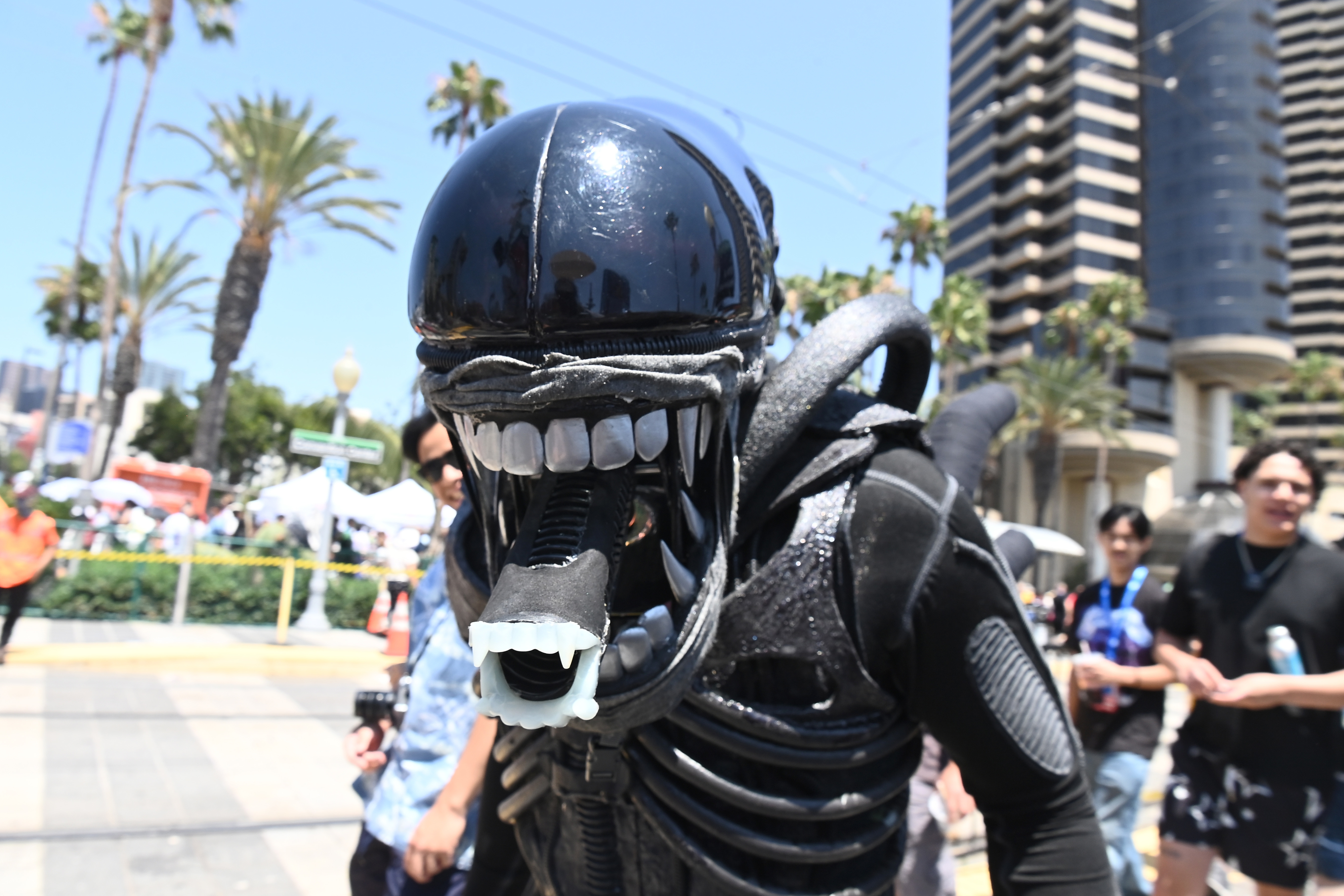 A person in a detailed, creature-like costume with a large mouthpiece walks outdoors at a crowded event. Palm trees and tall buildings are in the background