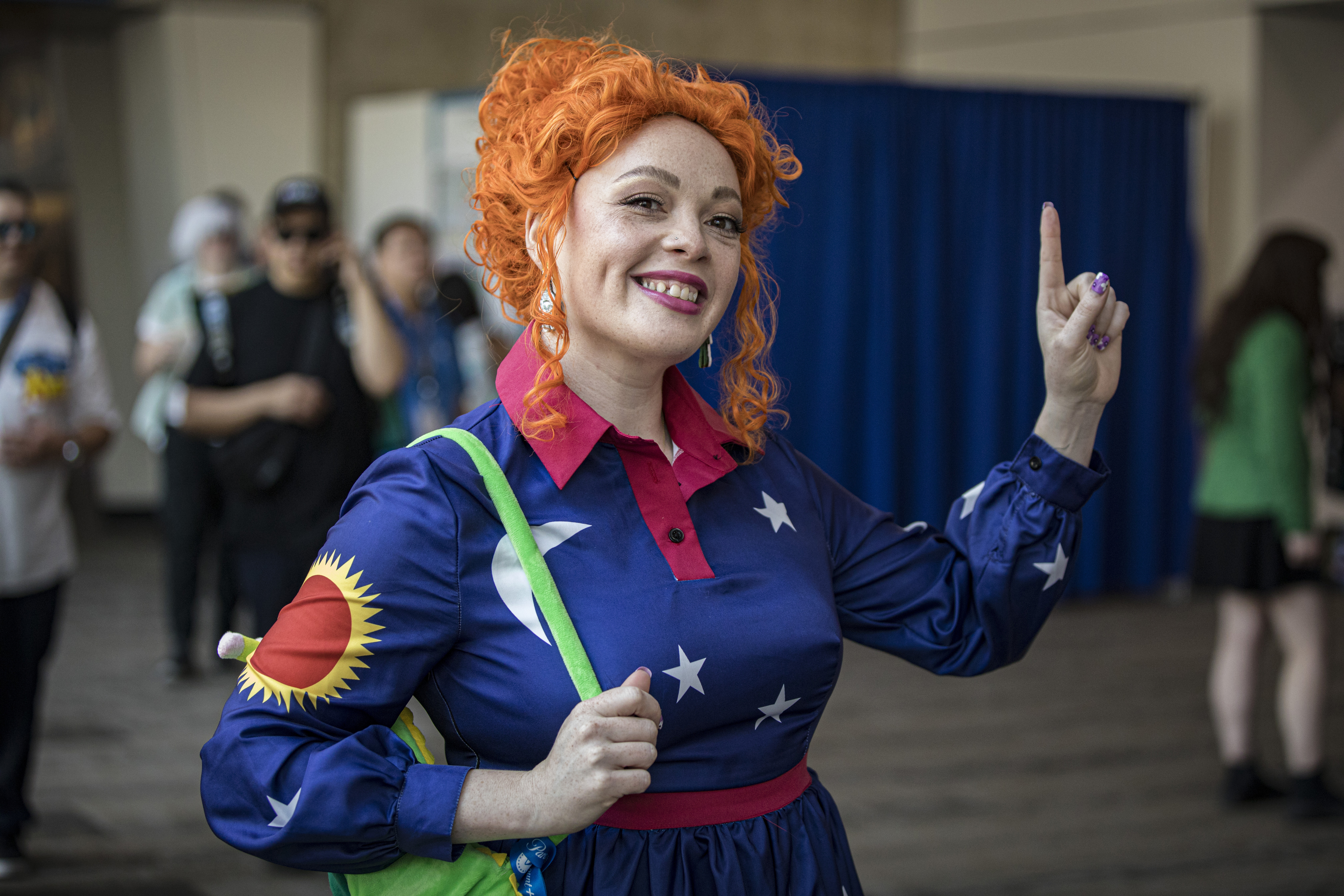 A person dressed as Ms. Frizzle from "The Magic School Bus" smiles and points upwards, wearing a dress with sun, moon, and star patterns