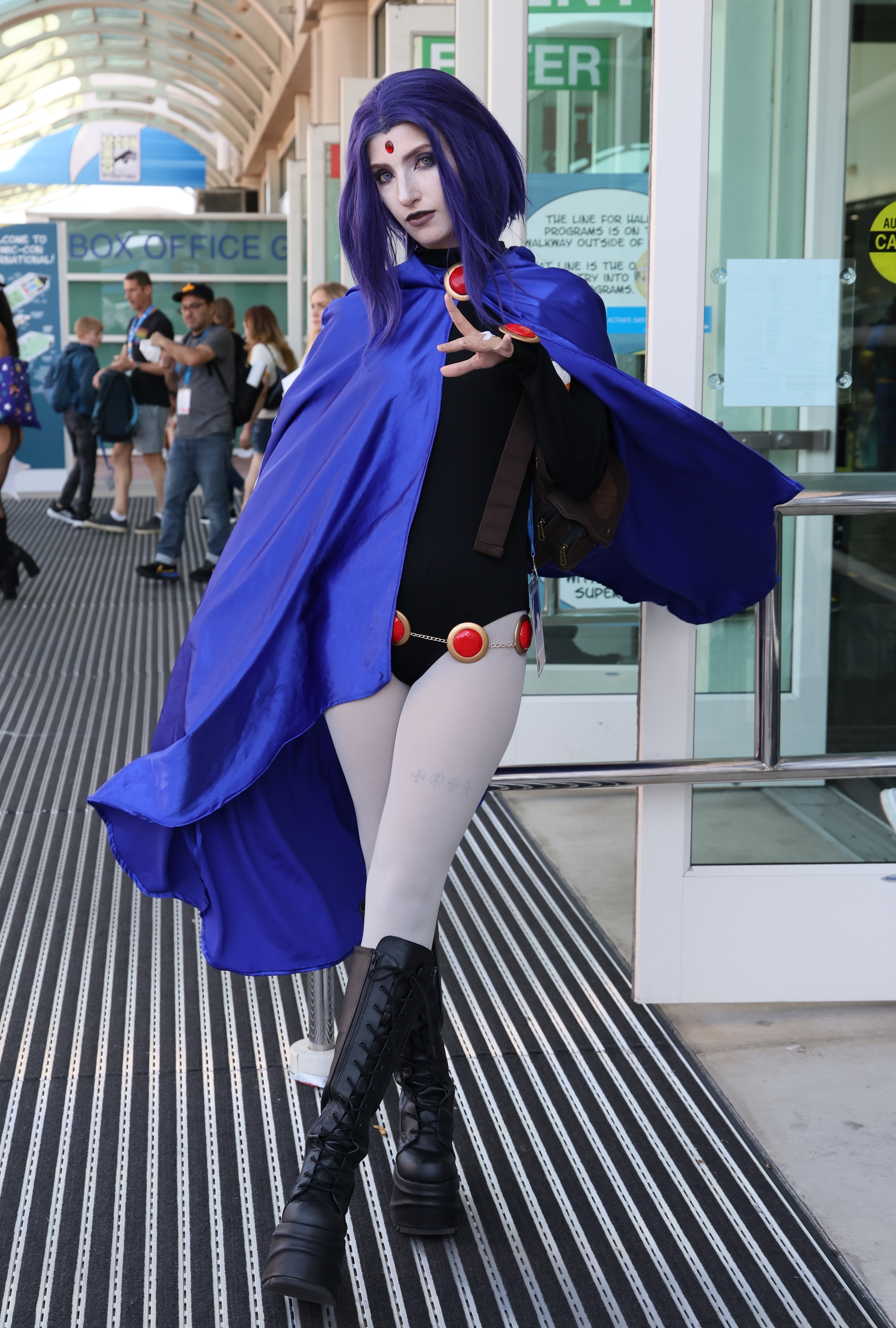 Raven cosplay from Teen Titans, wearing a purple hooded cape, black top, grey tights, and black boots. Background shows a busy convention area