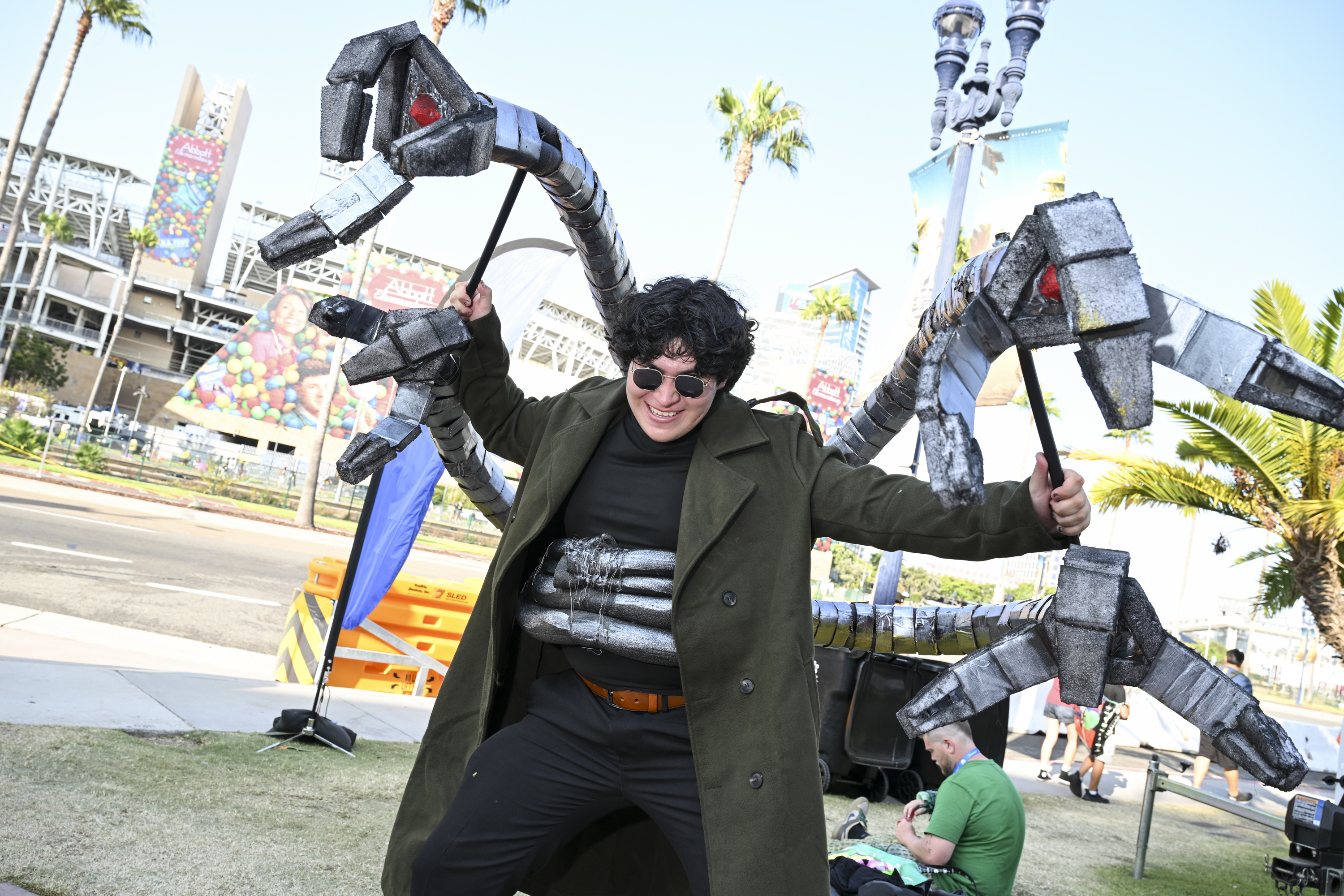 Person dressed as Doctor Octopus from Spider-Man, wearing a long coat and mechanical arms, poses at an outdoor event with palm trees and people in the background