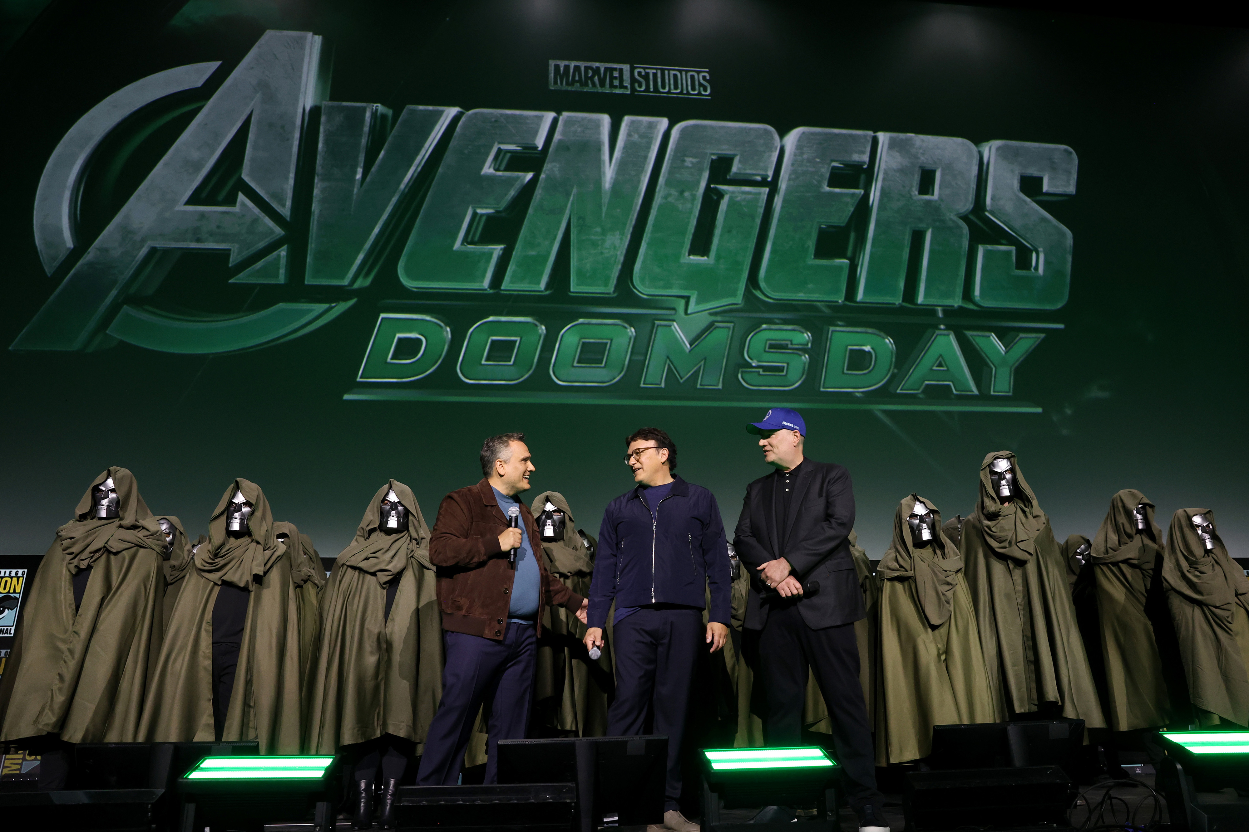 The Russo brothers and Kevin Feige stand in front of a large &quot;Avengers: Doomsday&quot; sign, flanked by people in hoods