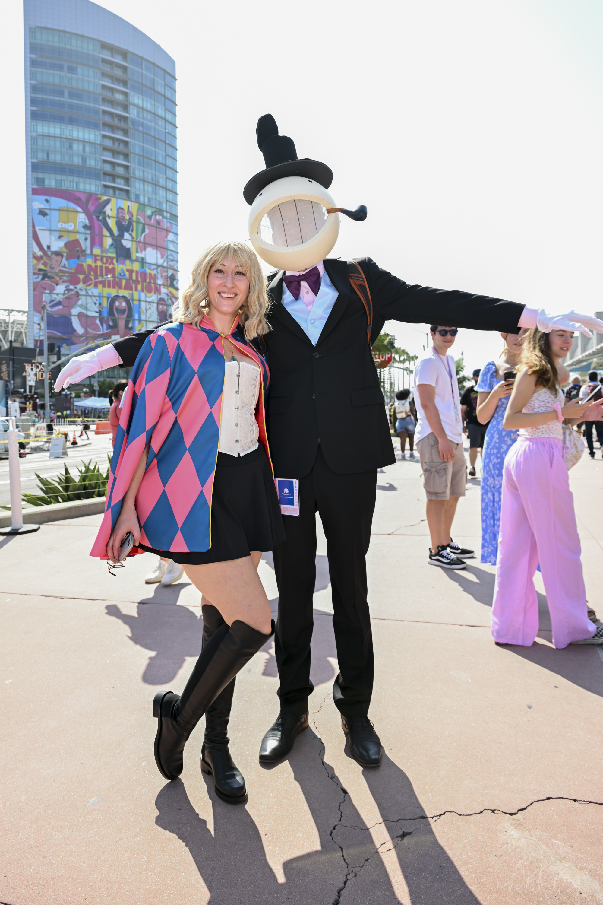 People in vibrant cosplay outfits, including an adventurous costume with a cape and a whimsical character with a round mask, outside a convention center