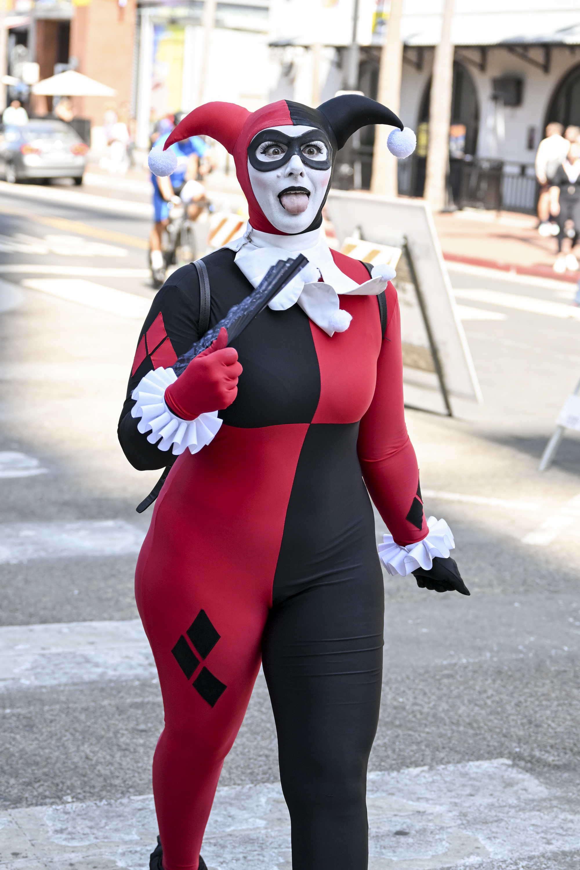Person dressed as Harley Quinn walking on the street, holding a prop hammer and sticking out their tongue