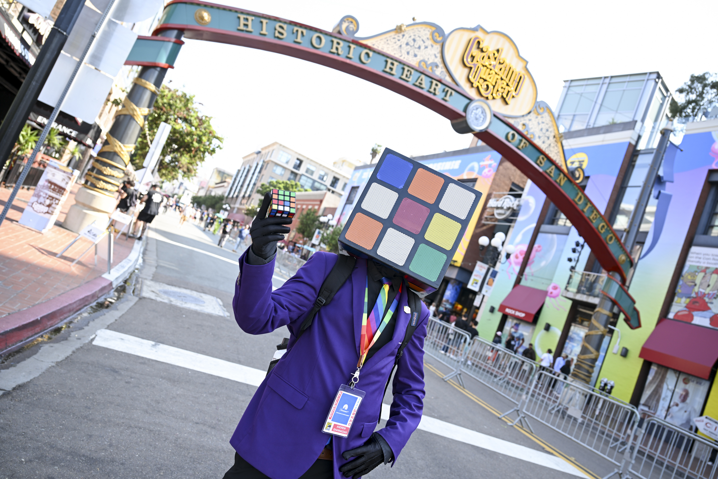 Person in a purple jacket with a Rubik's Cube head, holding a Rubik's Cube. They stand under the "Historic Heart of San Diego" arch