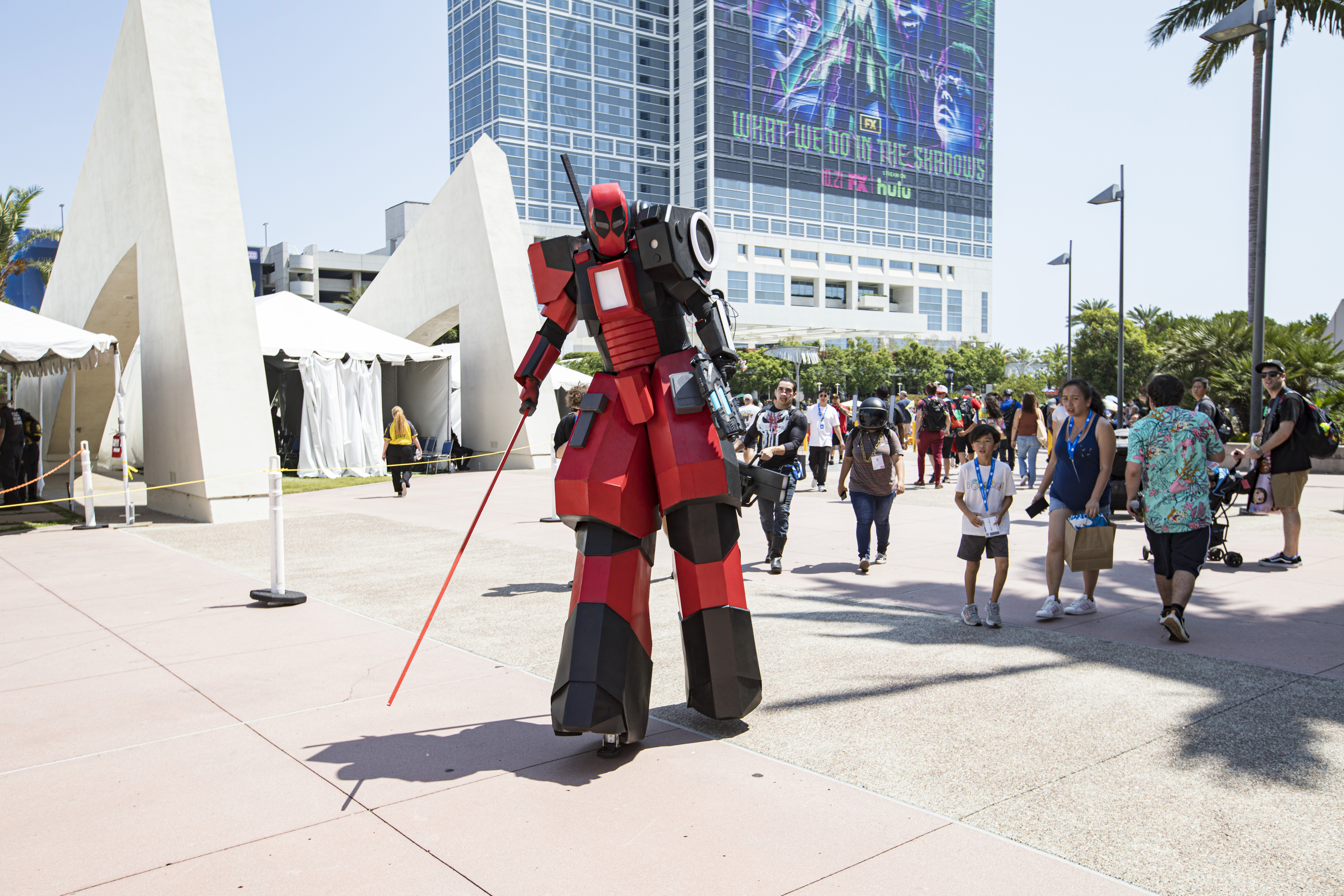A person in a tall, red and black mech costume walks outdoors at a crowded event, likely a convention, with a futuristic building in the background