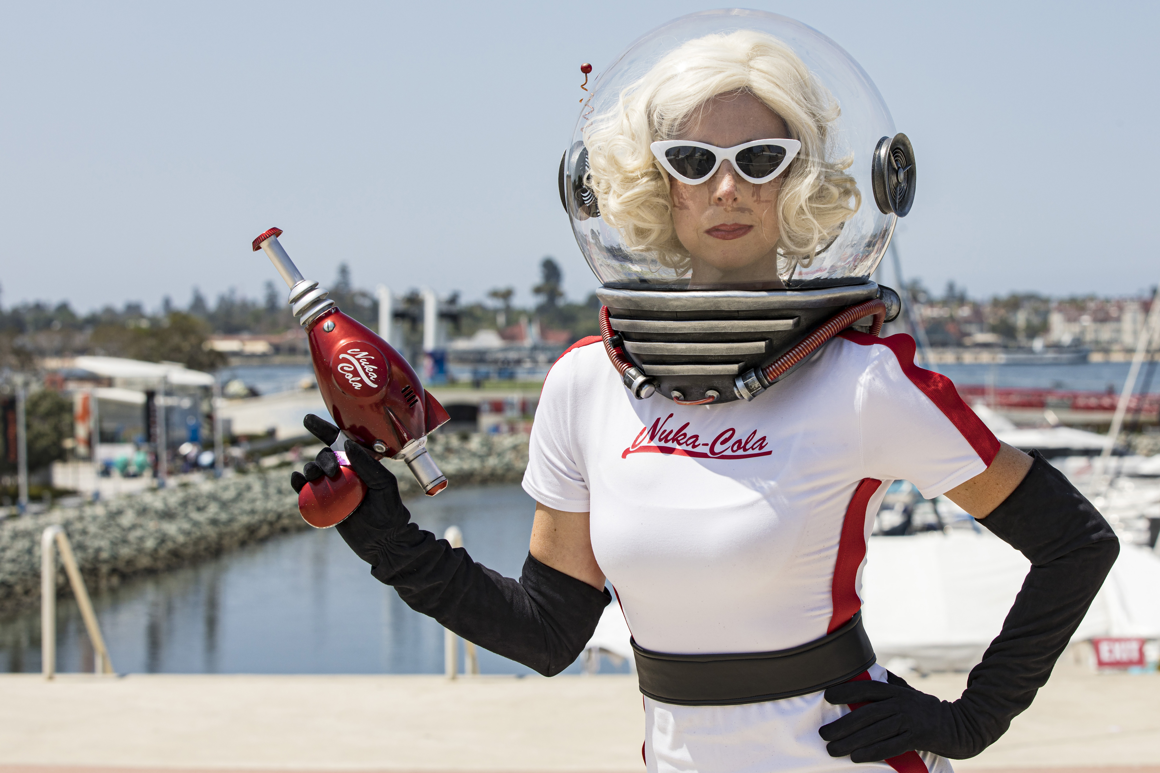 Person in a "Nuka-Cola" spacesuit costume outdoors by water, holding a ray gun, wearing white wig, white sunglasses, and a clear helmet with black gloves
