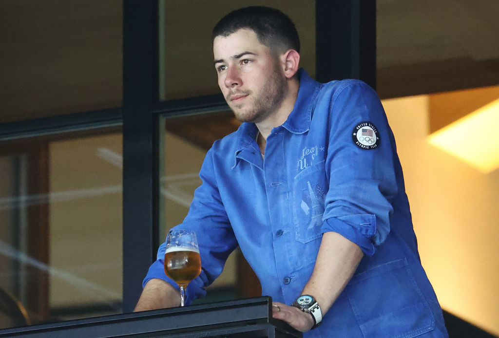 Nick Jonas, wearing a blue, long-sleeved shirt, stands on a balcony holding a glass of beer
