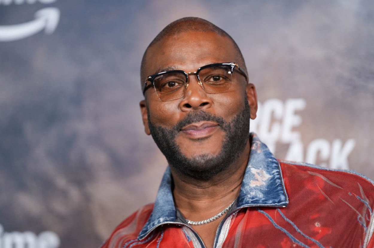 Tyler Perry is wearing stylish glasses and a red, denim-accented jacket. He is attending a public event or premiere