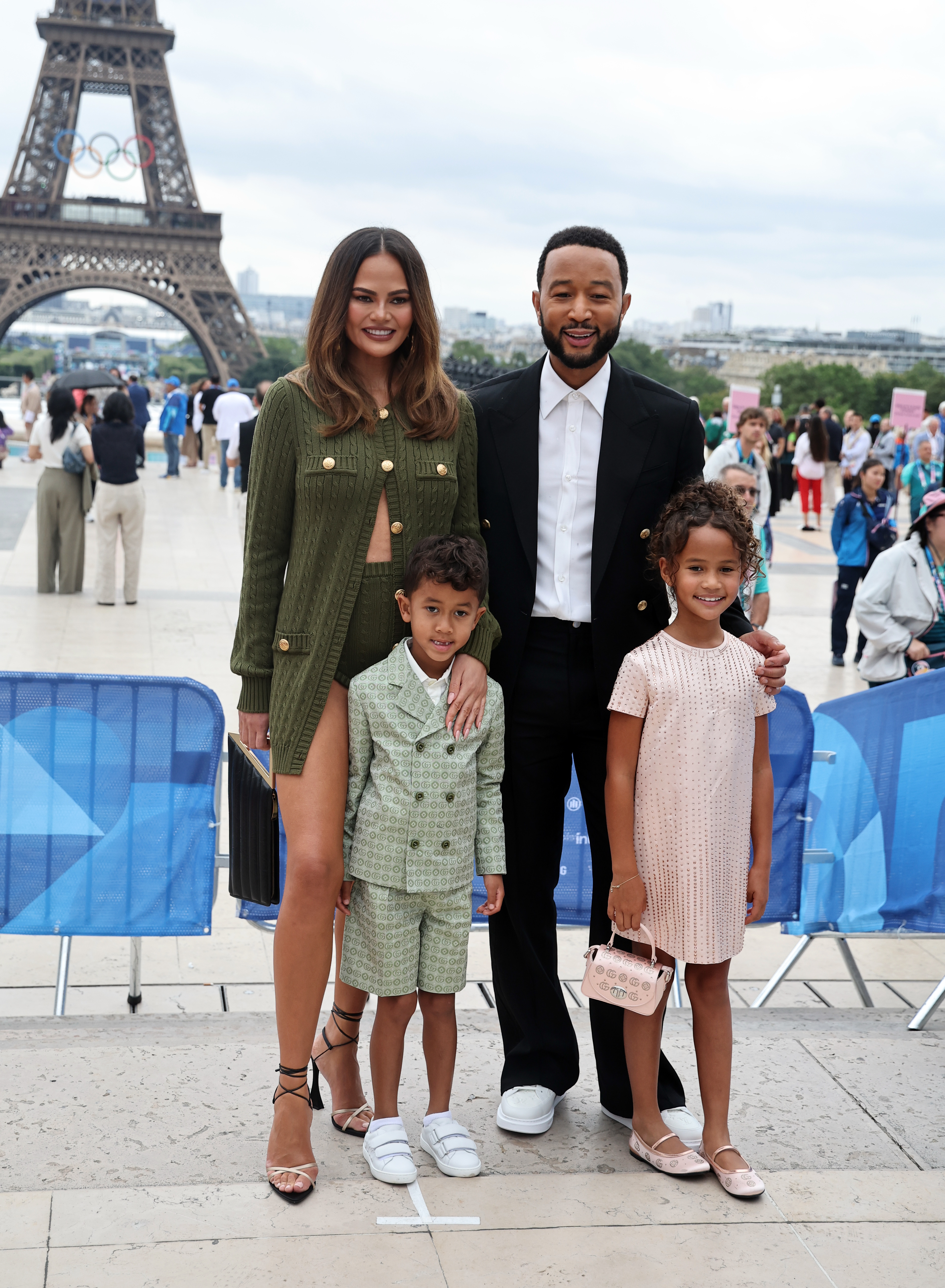Chrissy Teigen, John Legend, and their two children pose in front of the Eiffel Tower. Chrissy wears a green coat, John a black suit, and their kids in stylish outfits