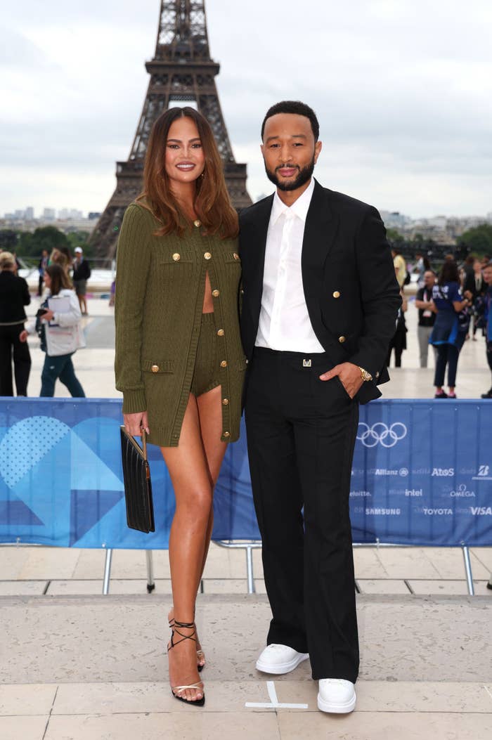 Chrissy Teigen and John Legend pose in front of the Eiffel Tower. Chrissy is wearing a stylish cardigan and shorts outfit. John is in a black suit with a white shirt
