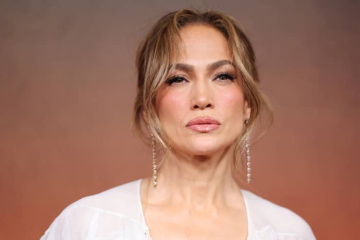 Jennifer Lopez wearing a white blouse and long earrings, with her hair styled in a loose updo, against a simple background