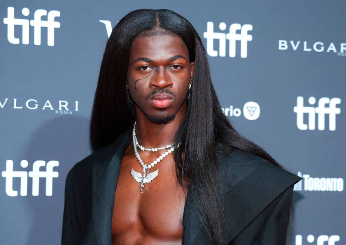Lil Nas X at the TIFF red carpet, wearing a black open blazer, layered pearl necklaces, and a facial tattoo near his eye