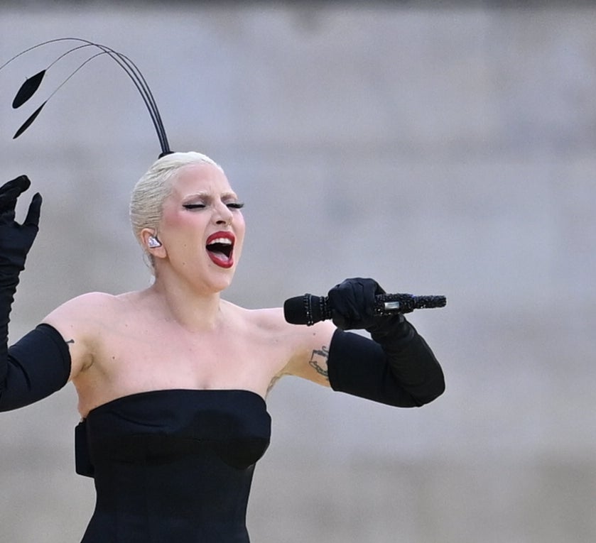 Lady Gaga performs on stage, wearing a strapless dark dress and matching gloves, with a dramatic headpiece featuring black leaves