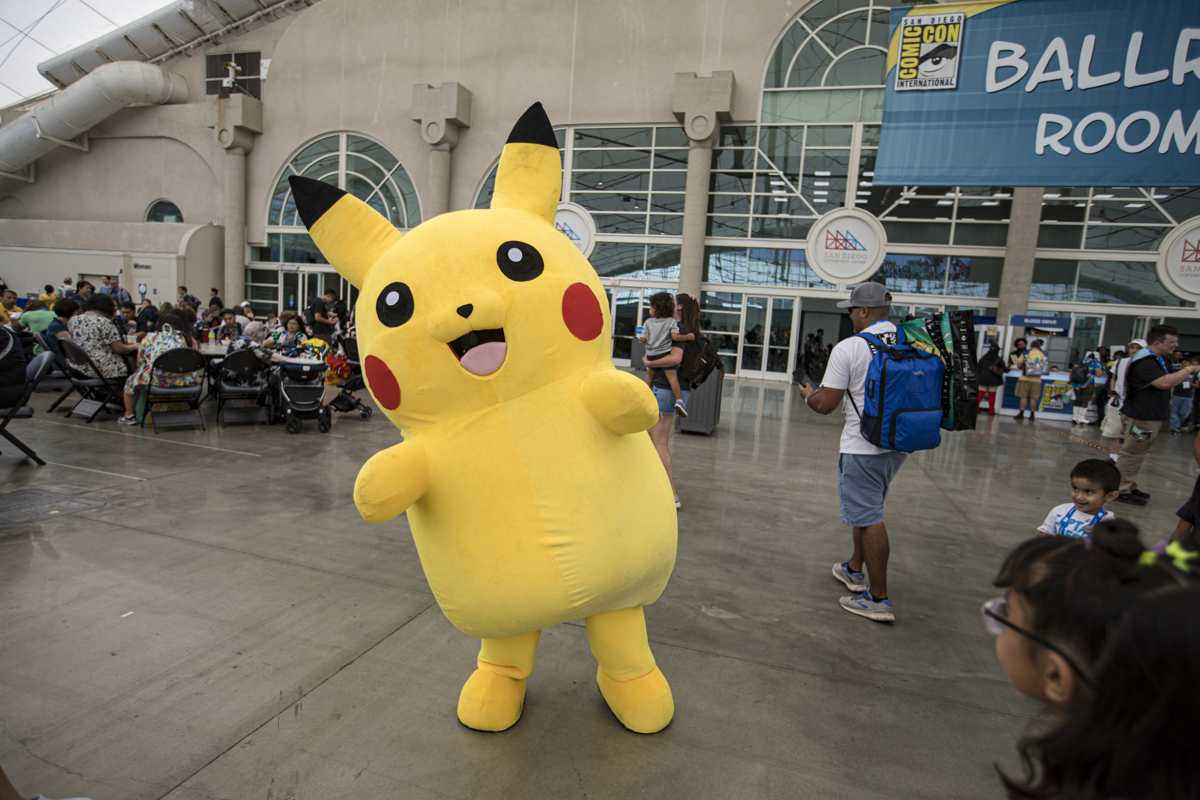 Pikachu mascot at San Diego Comic-Con posing inside a convention center, surrounded by attendees and a "Ballroom 20" sign in the background