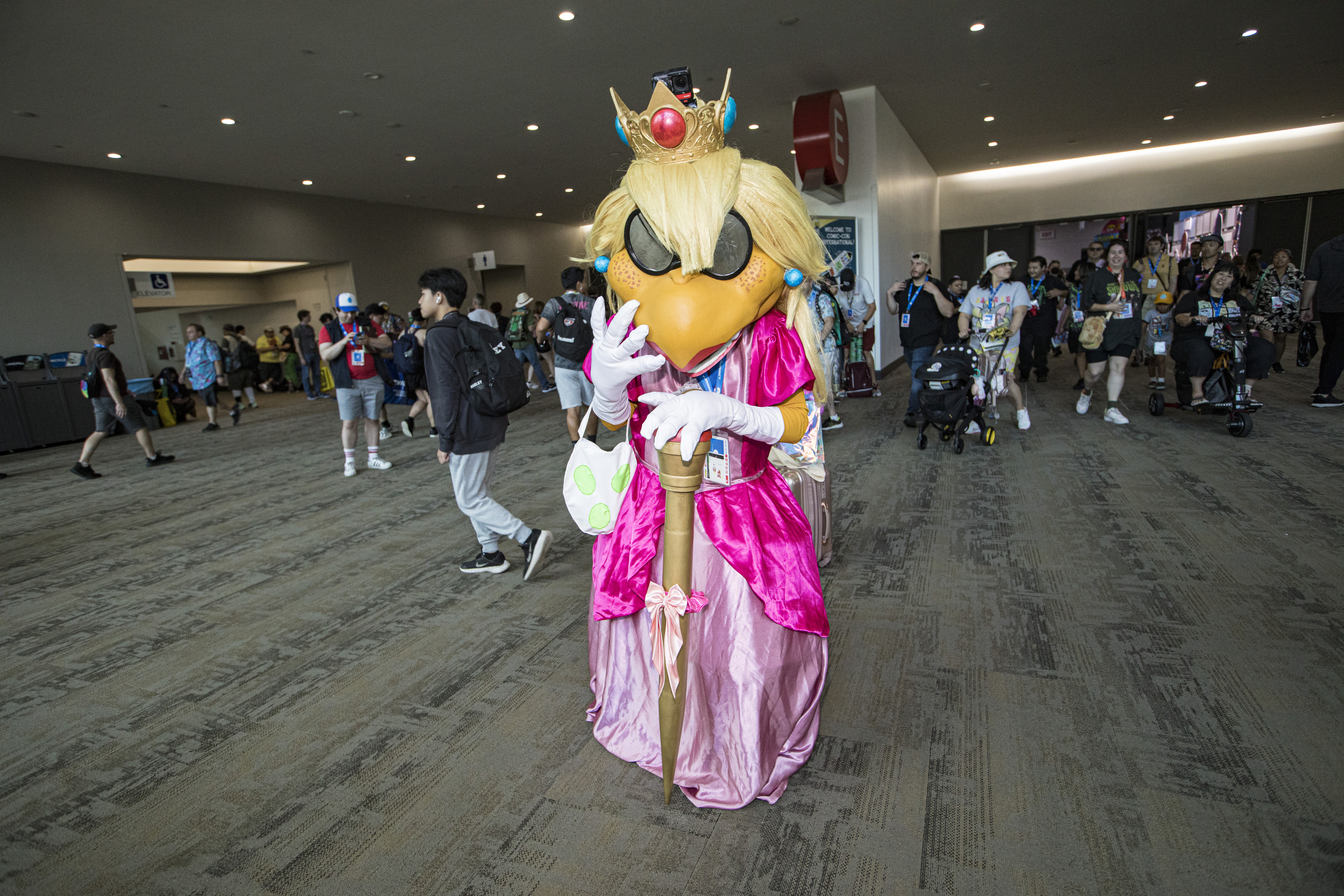 Person in a detailed Princess Peach costume from Super Mario stands in a convention hall, holding a green and white egg prop. People and strollers are in the background