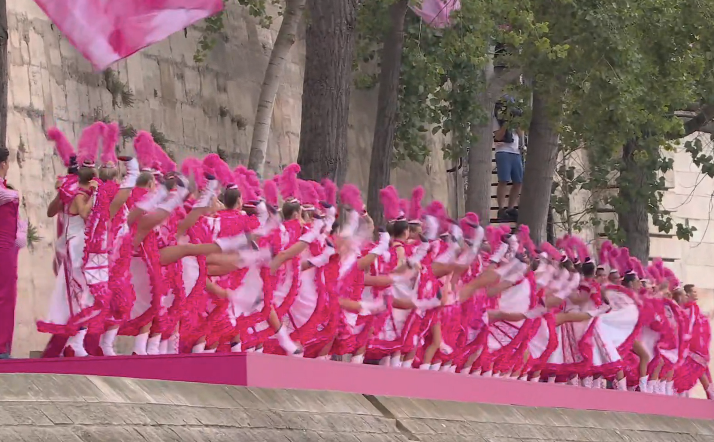 A synchronized group of dancers wearing matching red and white outfits performs an elaborate routine outdoors, with onlookers in the background