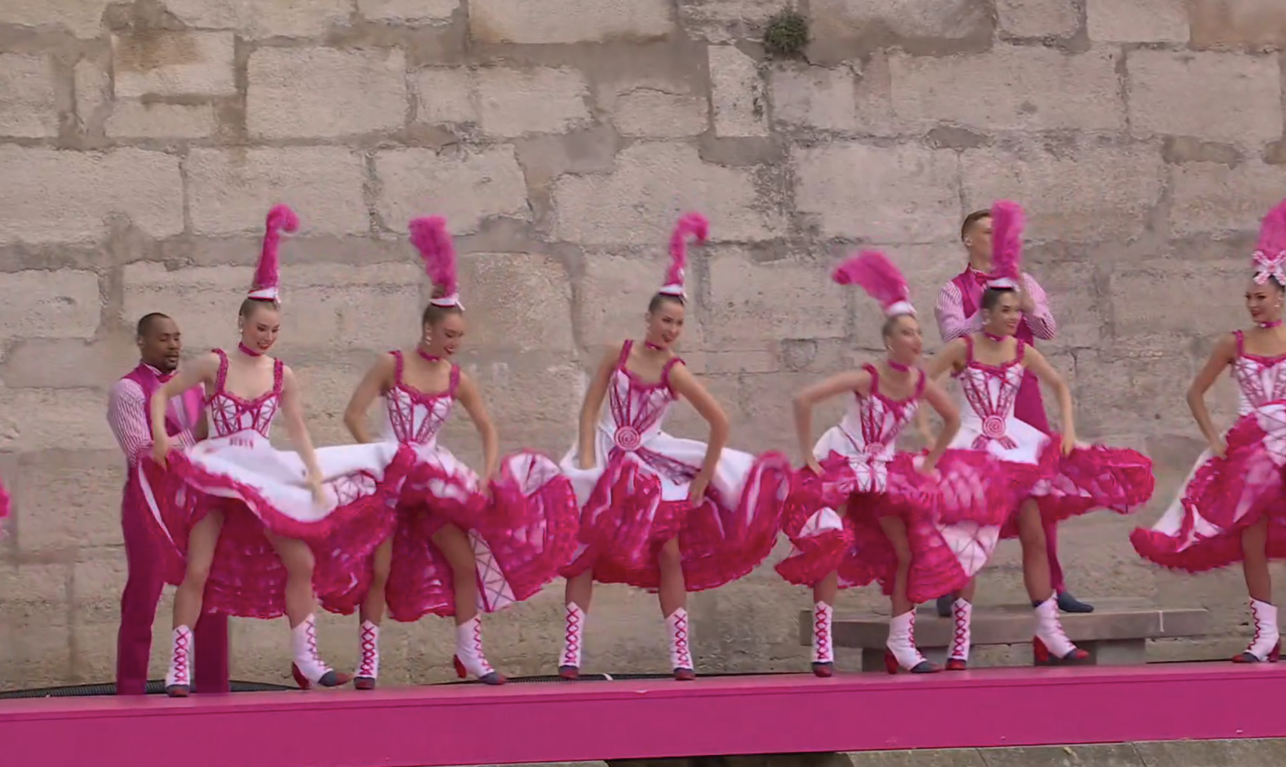 Group of Can-Can dancers performing on stage with a choreographer on the left, sporting elaborate high-kicking and ruffled skirts