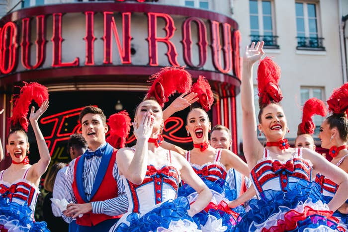 Performers in red, white, and blue costumes with feather headpieces pose in front of Moulin Rouge