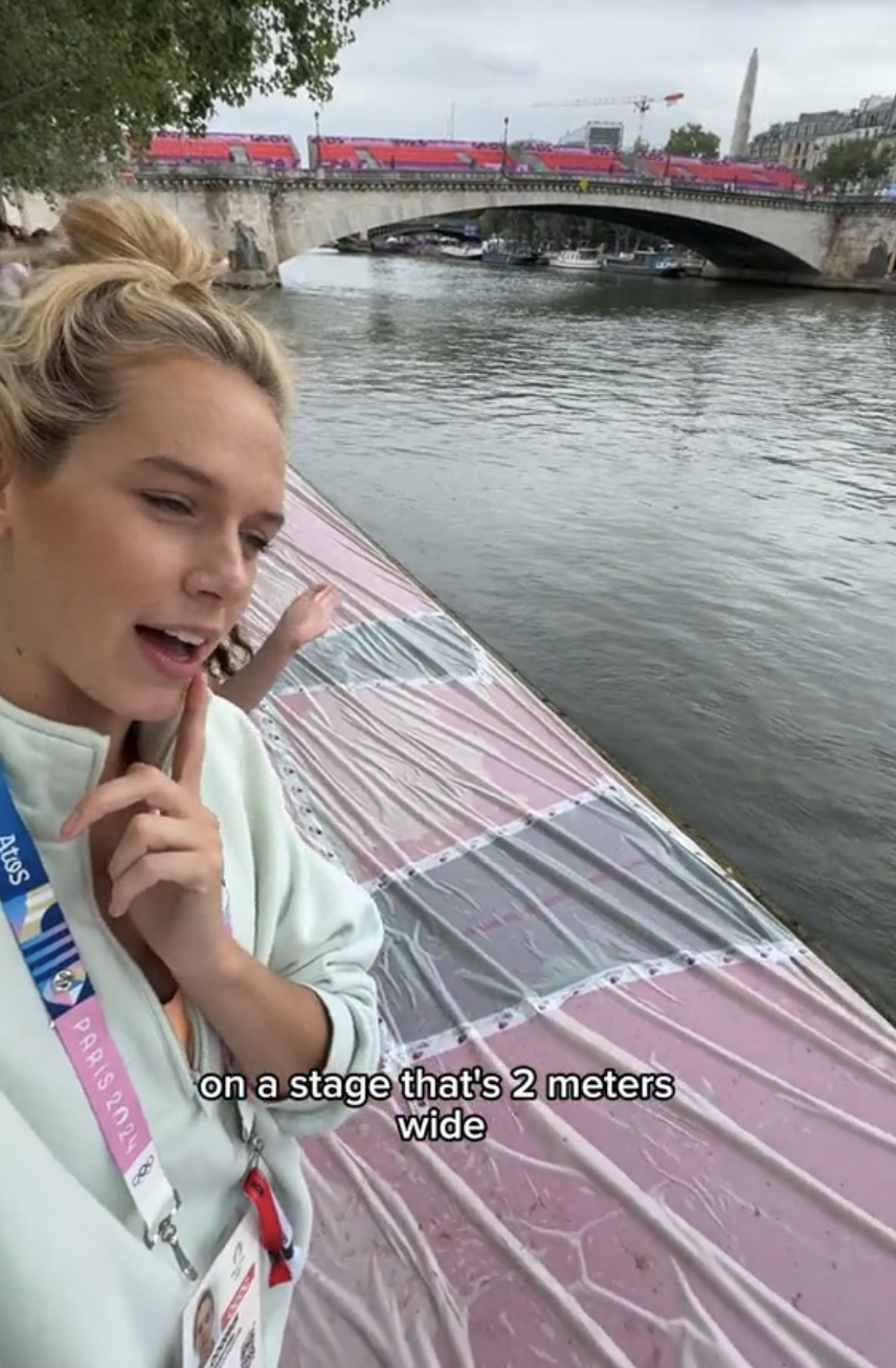 Person standing beside a river with a thinking pose, wearing a lanyard that says "Paris 2024." Text on the image reads, "on a stage that's 2 meters wide."