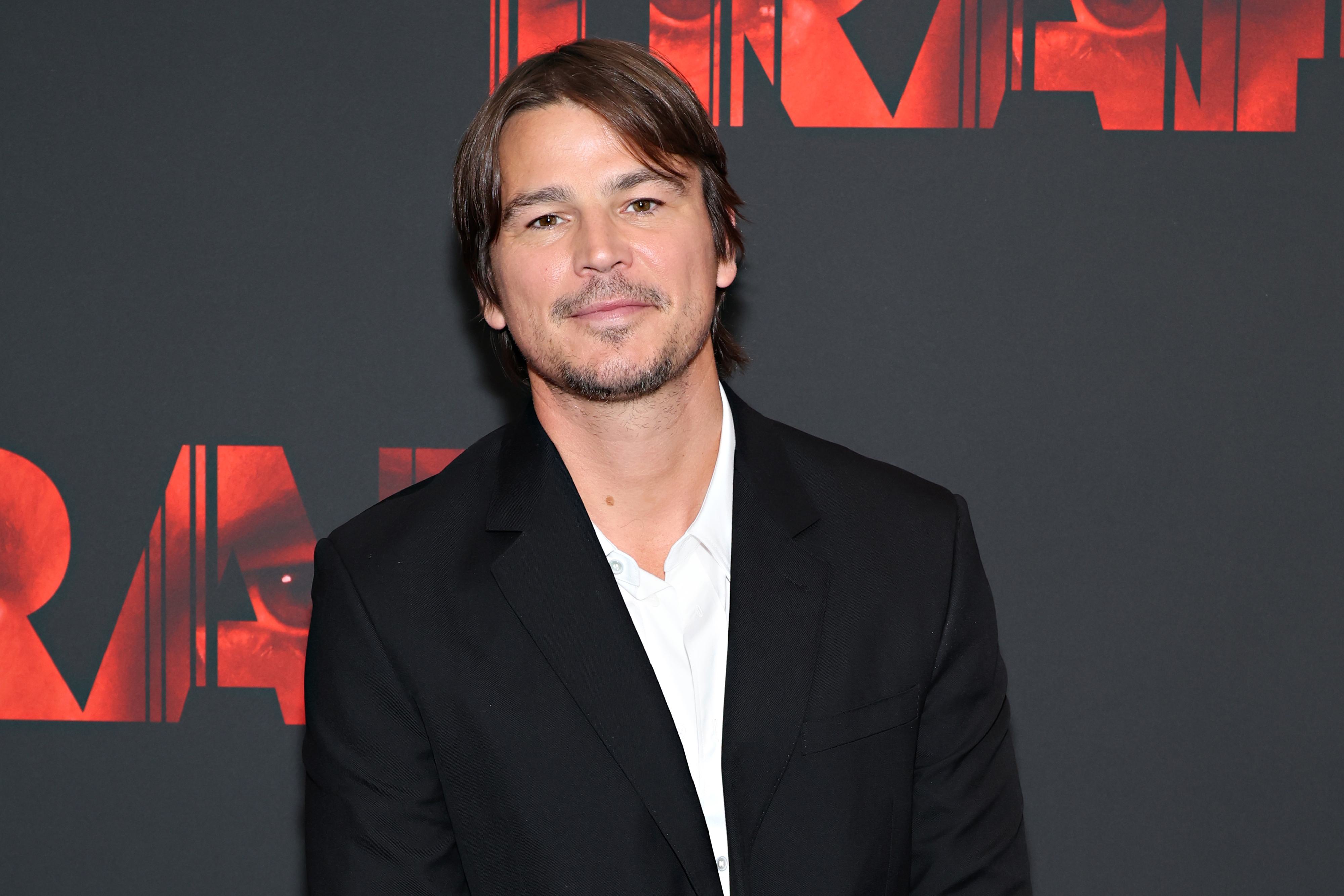 Josh Hartnett in a black suit jacket and white shirt, posing on a red carpet in front of a backdrop with red text