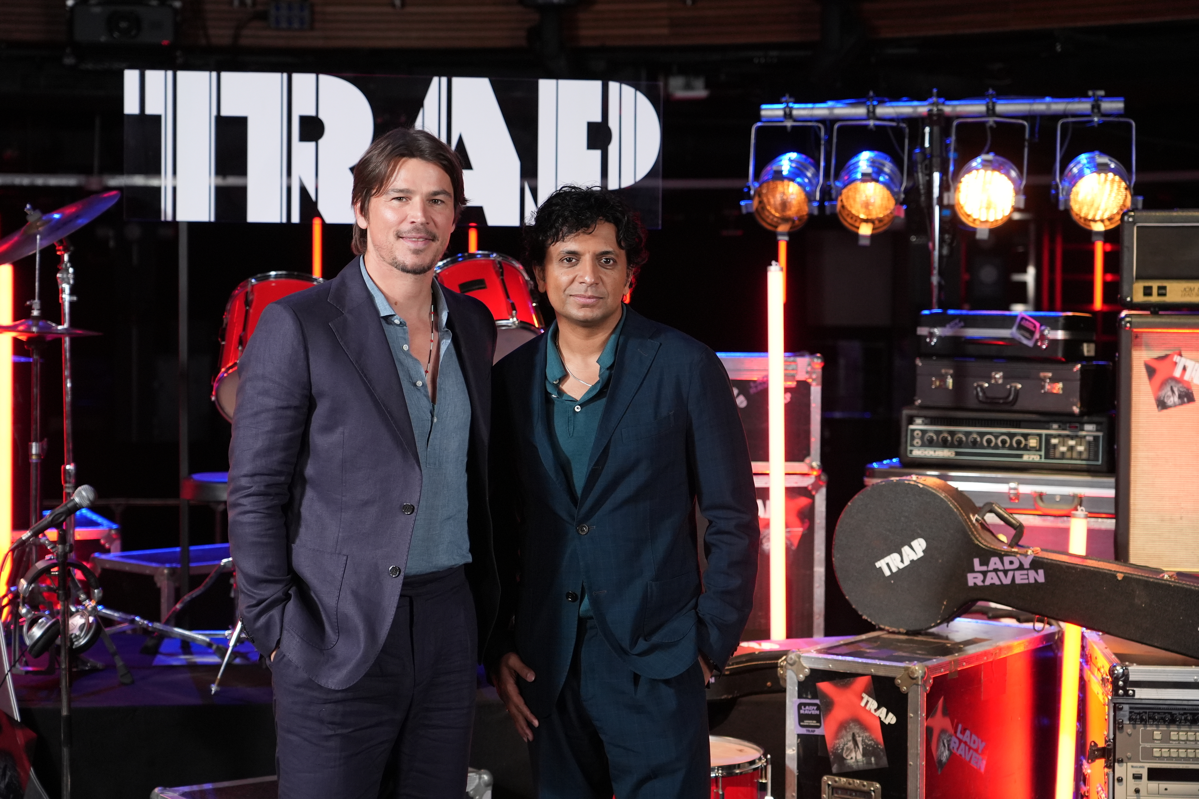 Josh Hartnett and M. Night Shyamalan on a music-themed set with drums, guitars, and various musical equipment in the background