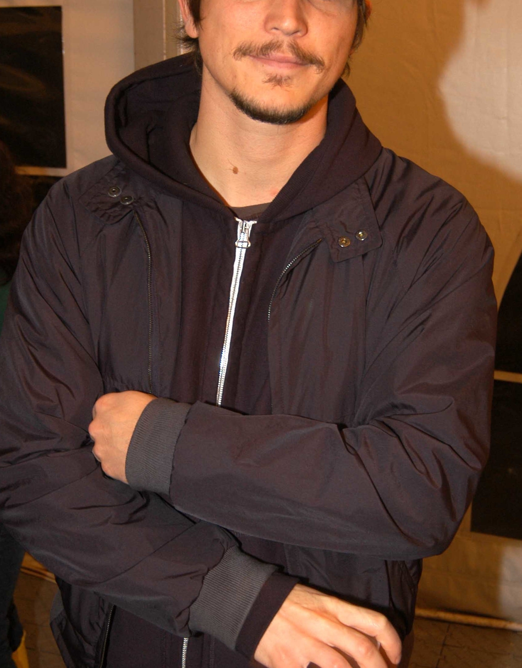 Josh Hartnett wearing a casual jacket, hoodie, and cap, with his arm crossed and holding a cup, poses for the camera. Another person in the background is partially visible