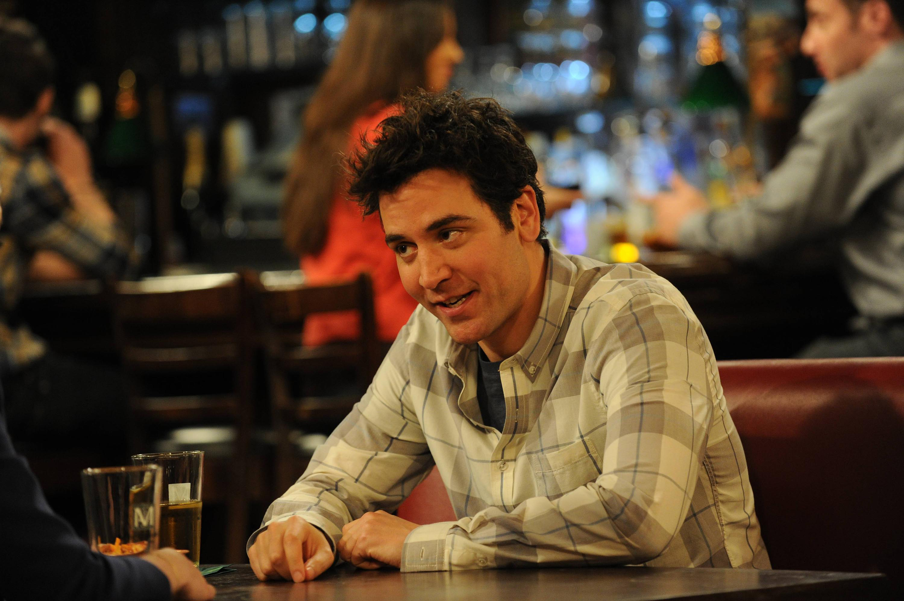 Josh Radnor sits at a bar booth engaged in conversation, holding a beverage in a scene from &quot;How I Met Your Mother.&quot;